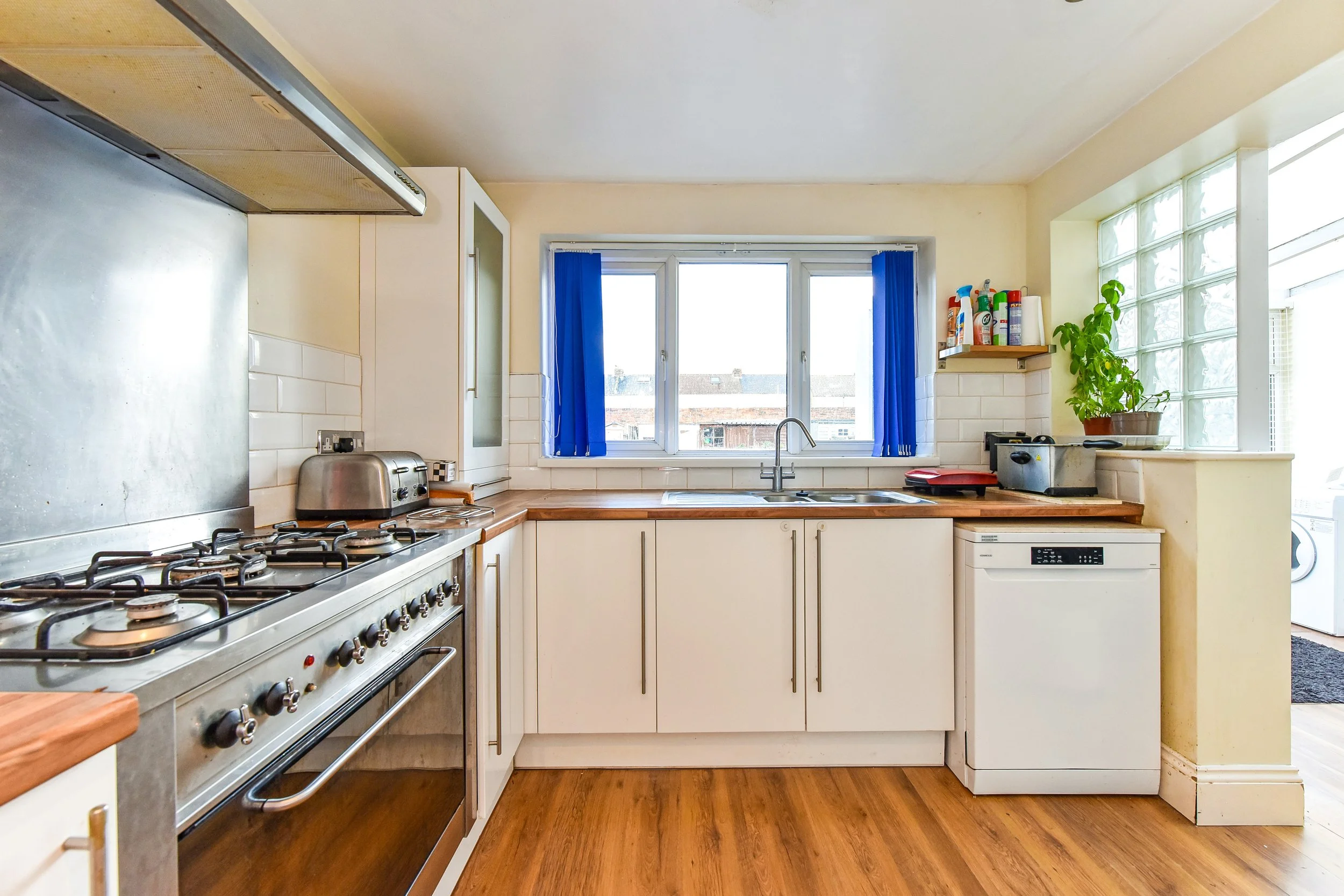 Kitchen with white cabinetry, wooden countertops, a gas stove, toaster, dishwasher, window with blue curtains, and a potted plant near a glass block wall.