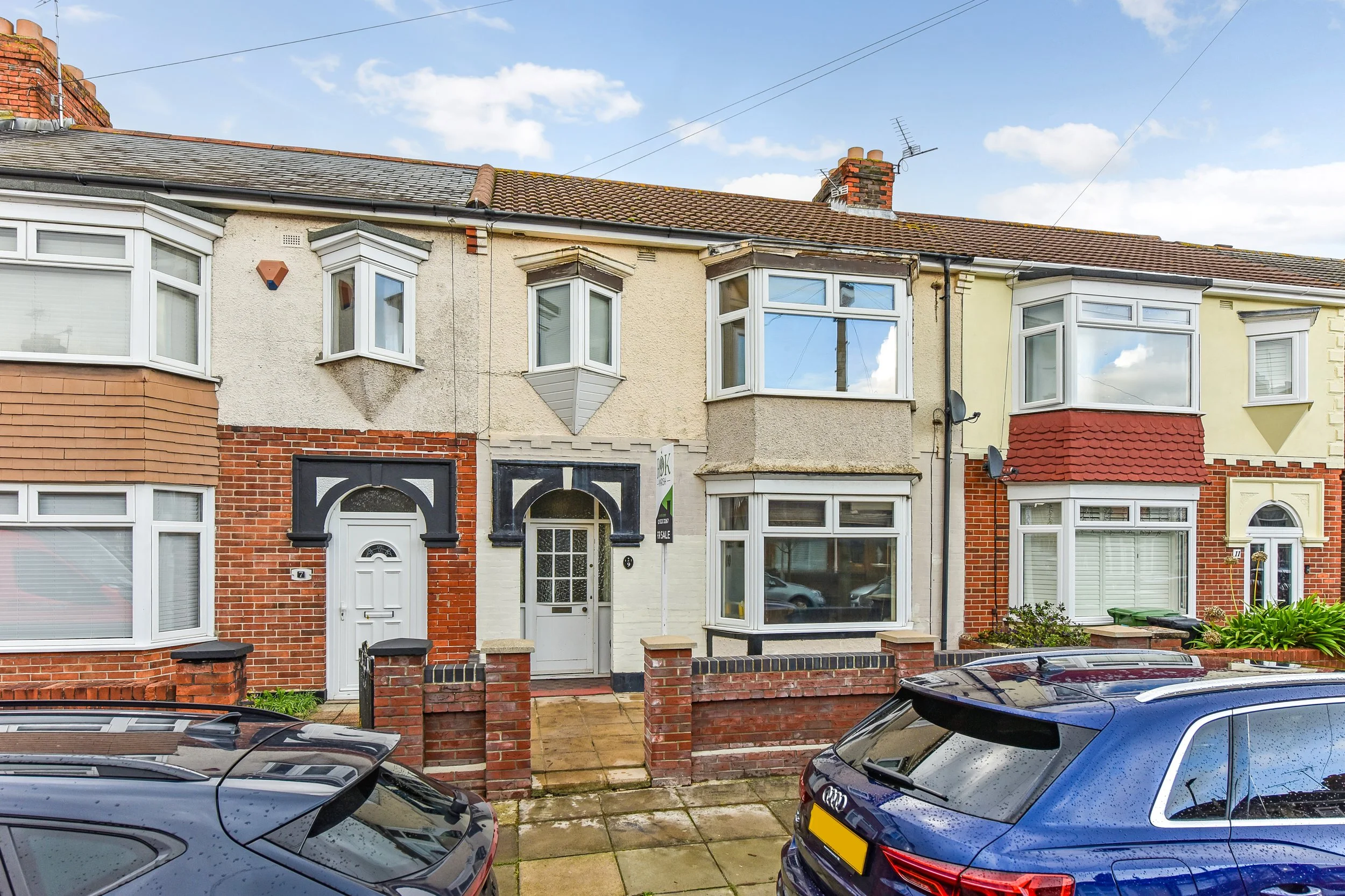 Row of terraced houses with brick and stucco facades, bay windows, and front yards, parked cars in front.