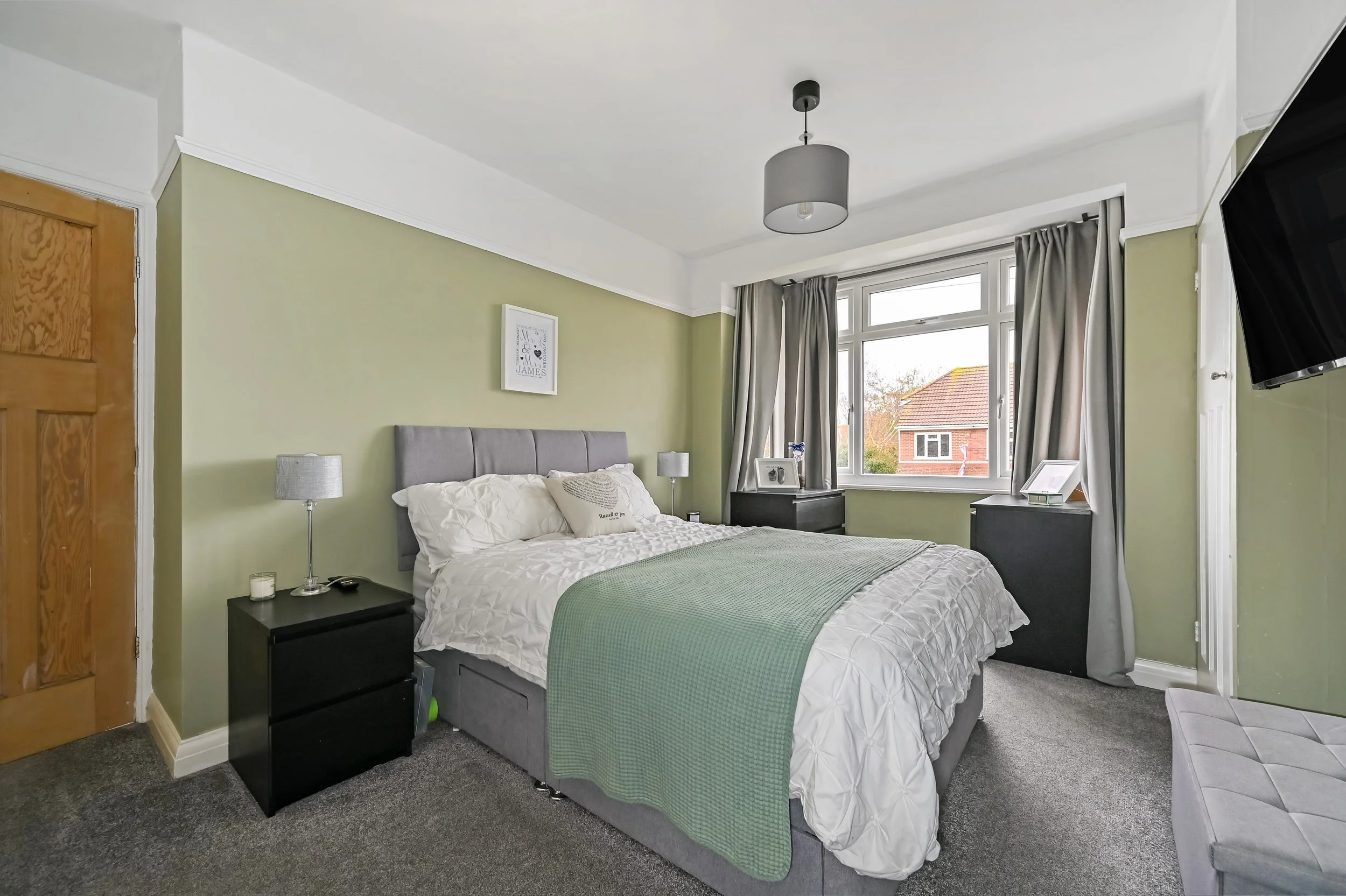 A bedroom with a bed, two black nightstands, a window with curtains, and a wall-mounted TV.