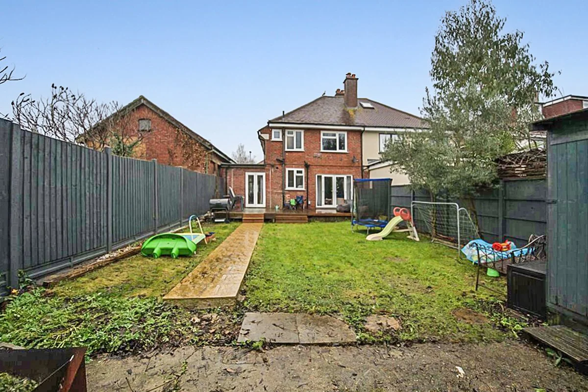 Backyard with a lawn, children's play equipment including a slide and tennis net, surrounded by a dark green fence, with houses in the background.