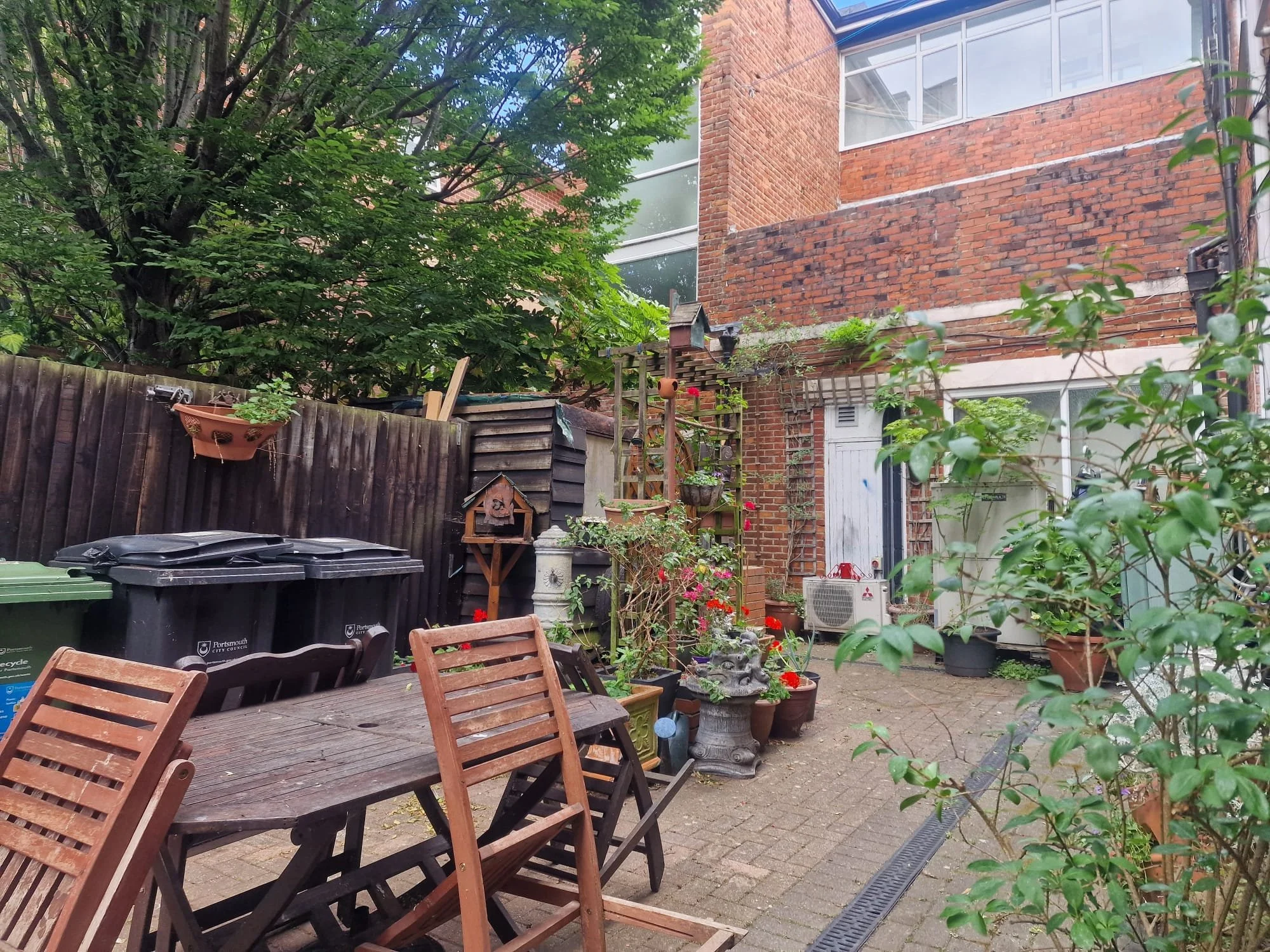 A backyard patio with a wooden table and chairs, potted plants, a wooden fence, and brick buildings in the background.