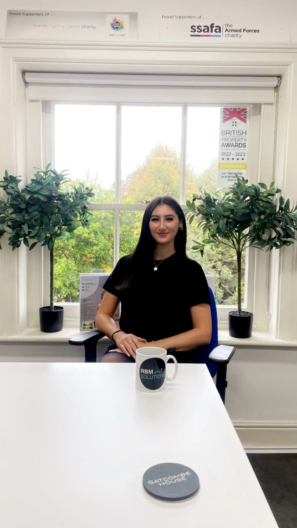 A young woman with long dark hair and a black shirt sitting at a white desk in an office with a large window behind her. The window has two potted plants on the windowsill and a view of trees outside. There are various posters and signs on the wall, including awards and charity information. On the desk, there is a mug with the logo 'RBM Solutions' and a round object with 'Gatcombe House' written on it.