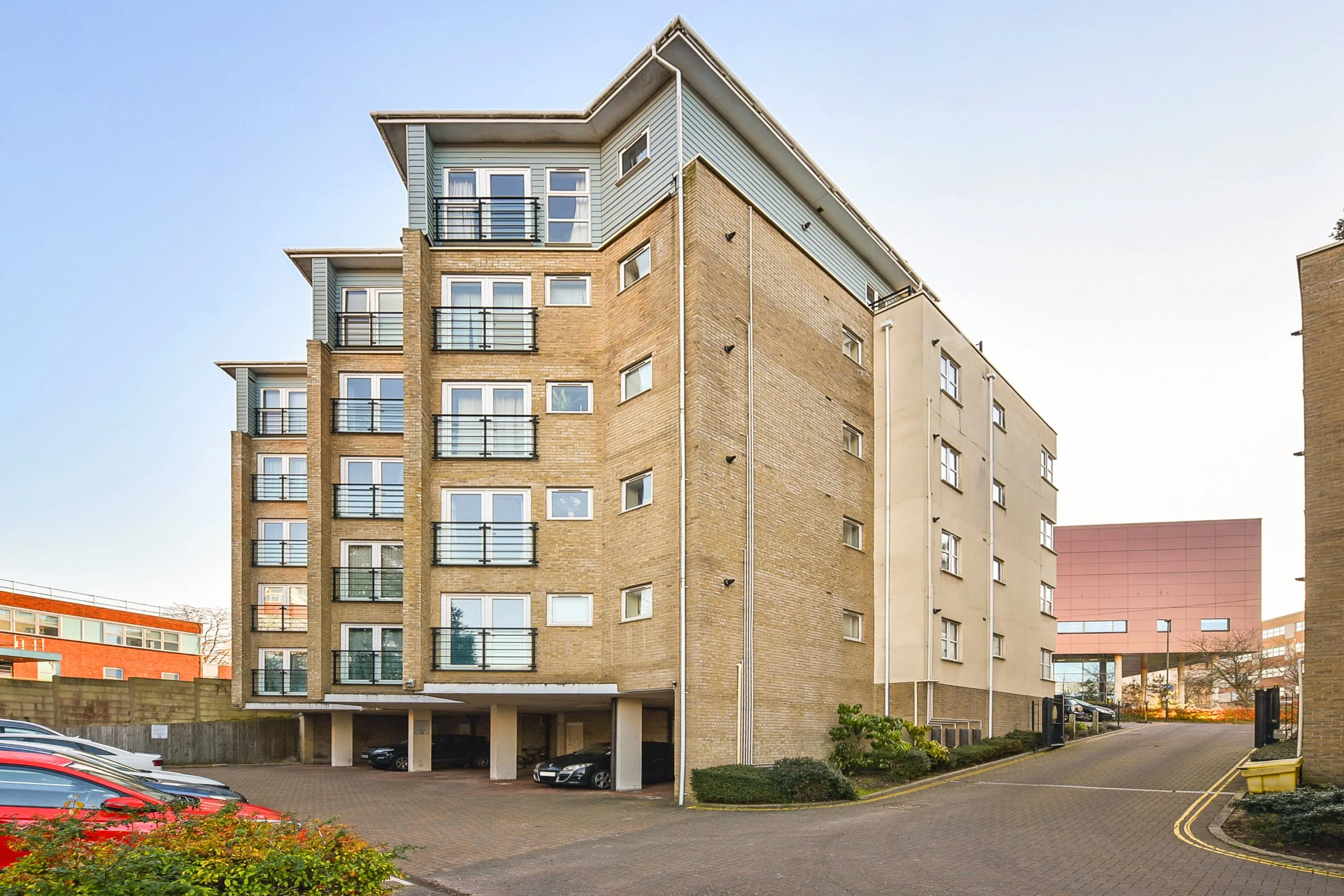 Multi-story residential apartment building with parking area underneath and a paved driveway leading to a gate, with modern and traditional architectural elements.