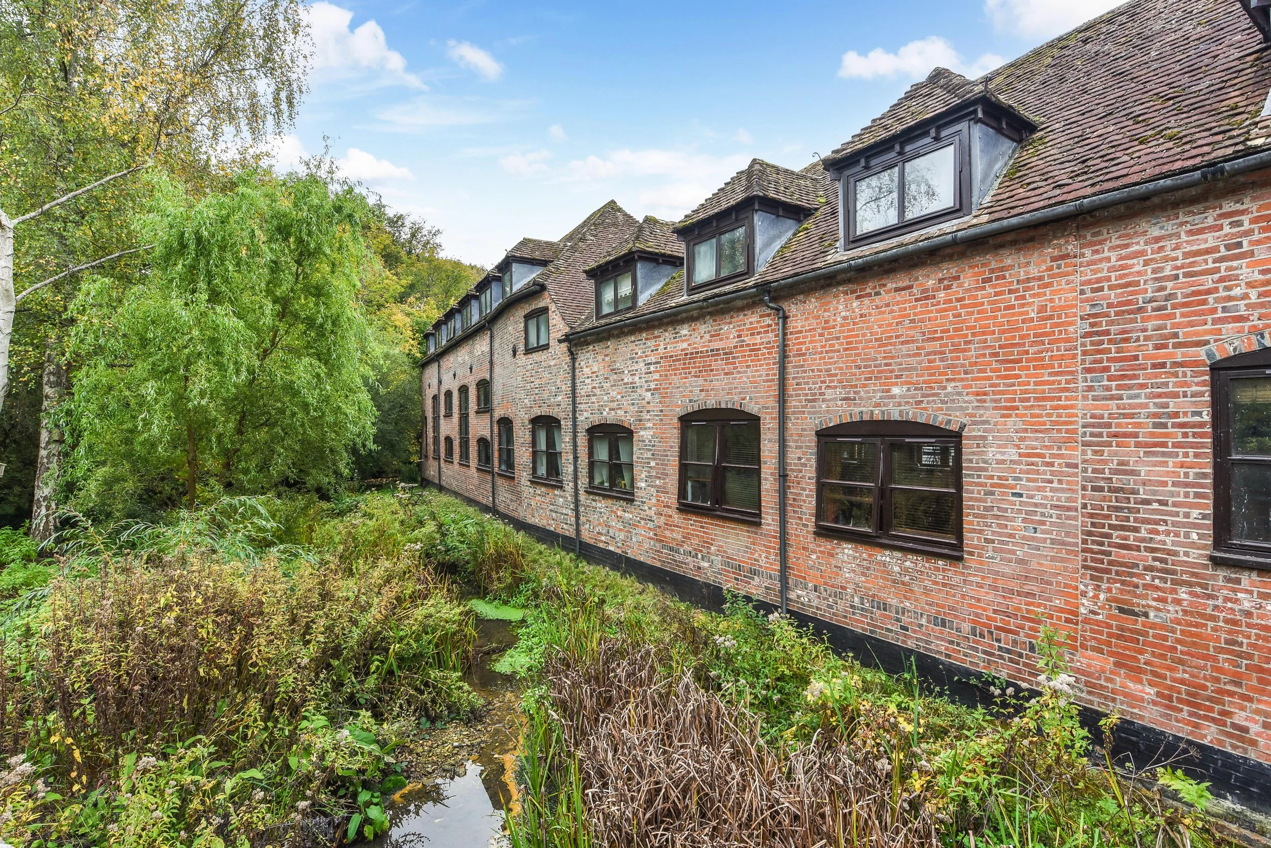 Brick apartment building with multiple windows near a small pond surrounded by green trees and plants.