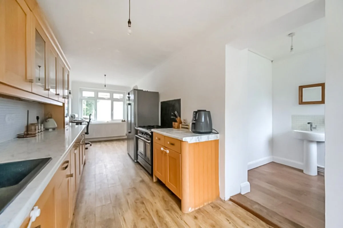 Kitchen with wooden cabinets, marble countertops, and wooden flooring, with a window at the end, a stove, and a small island with a coffee machine on top; an adjacent room with a sink and mirror.