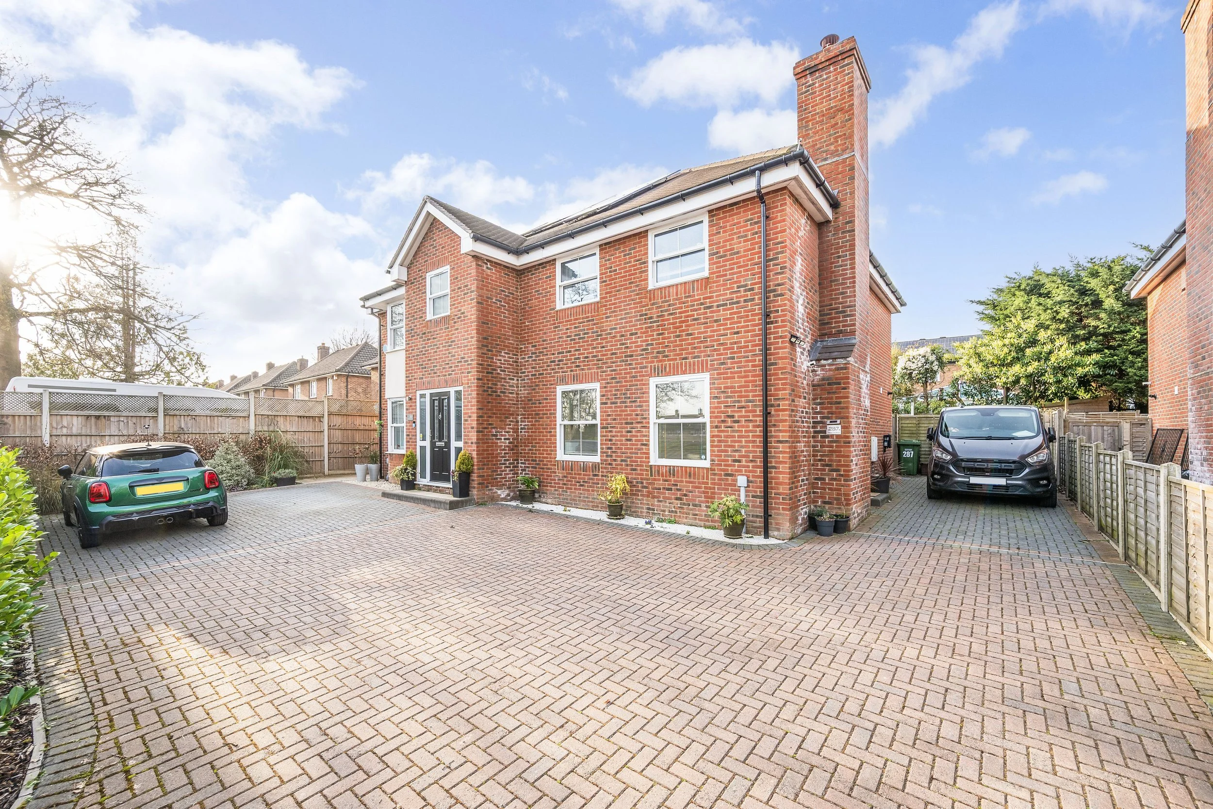 Brick residential house with a driveway, two cars parked, and potted plants outside on a sunny day.