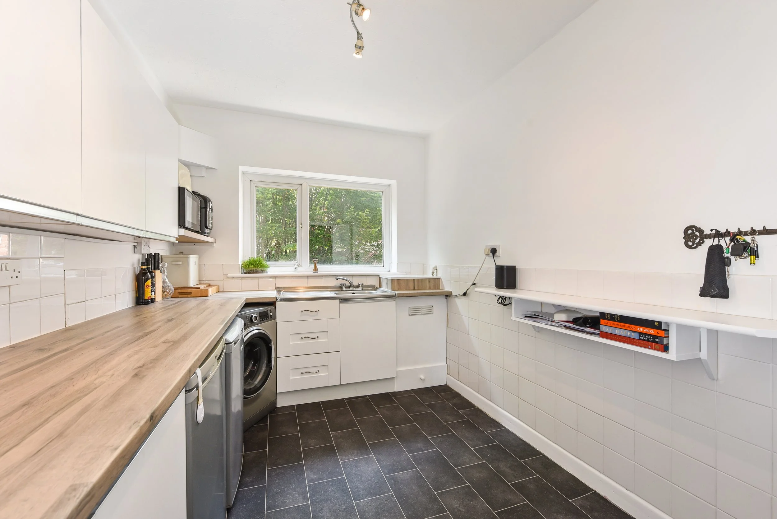 Kitchen with white cabinets, a wooden countertop, a washing machine, a window with greenery outside, and a small shelf with books.