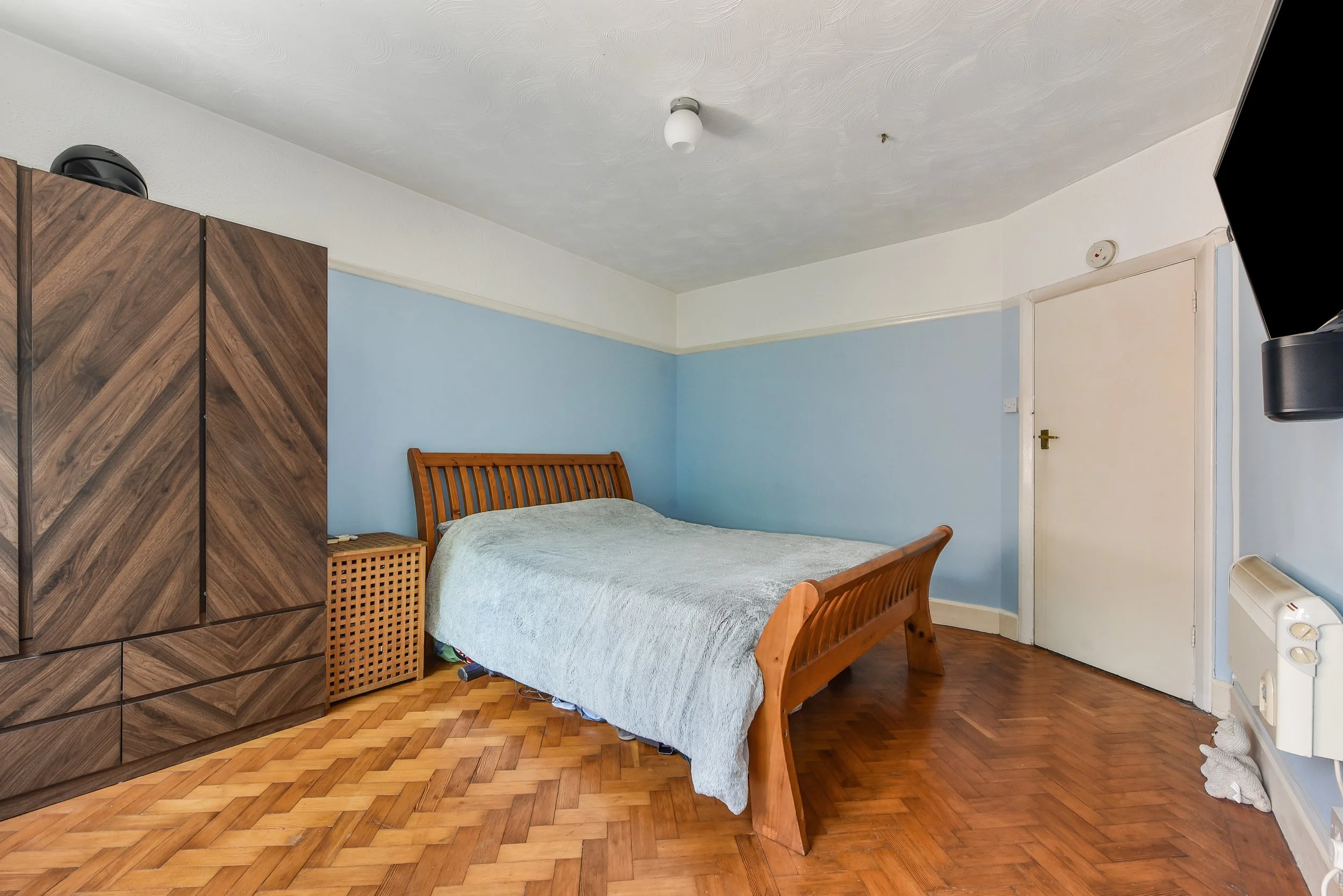 Bedroom with wooden bed, blue walls, wooden wardrobe, small bedside table, hardwood floor, white door, radiator, and plush toy on the floor near radiator.