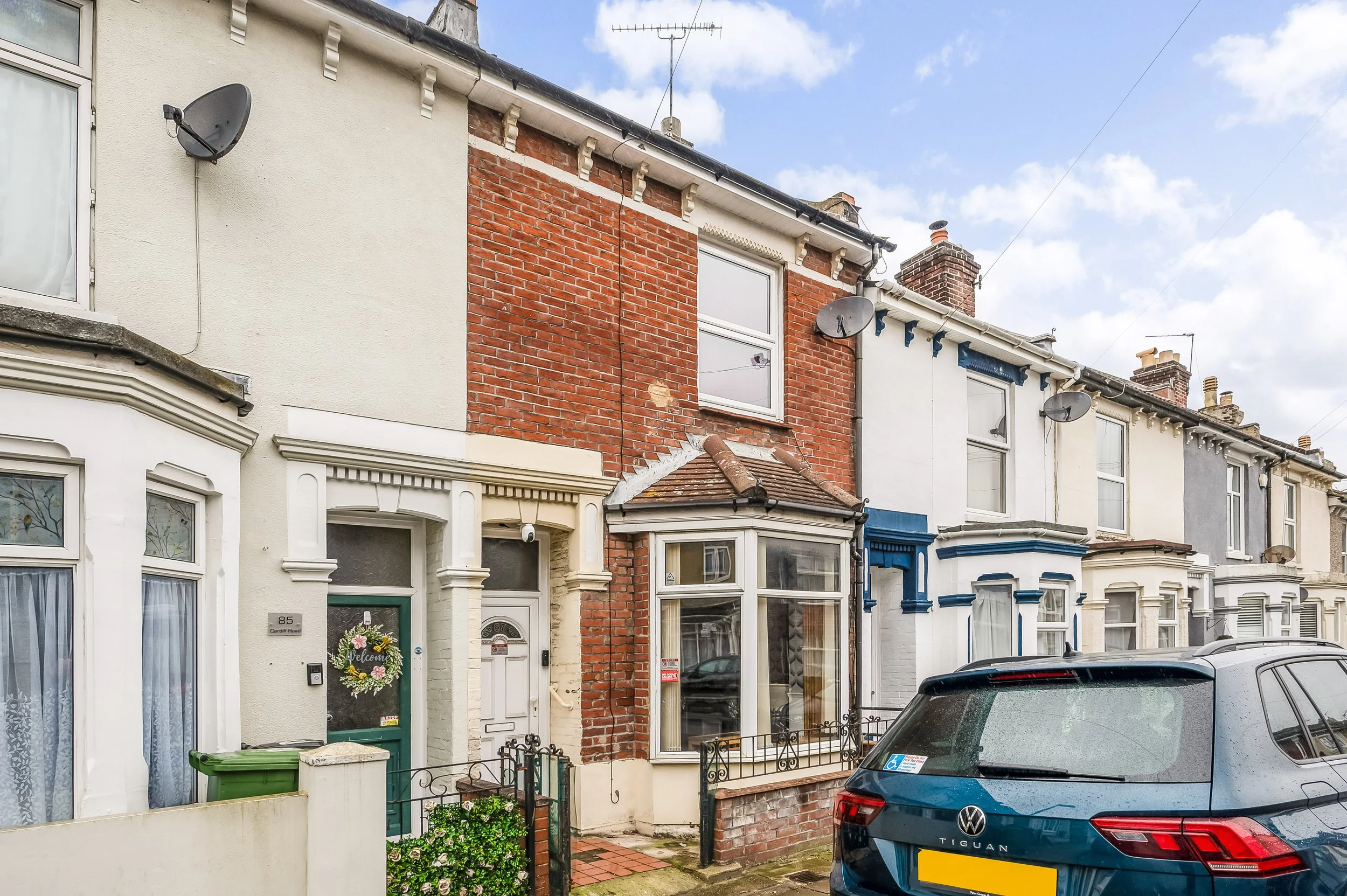 Row of terraced houses with brick and painted facades, some with bay windows, cars parked on the street, and a partly cloudy sky above.