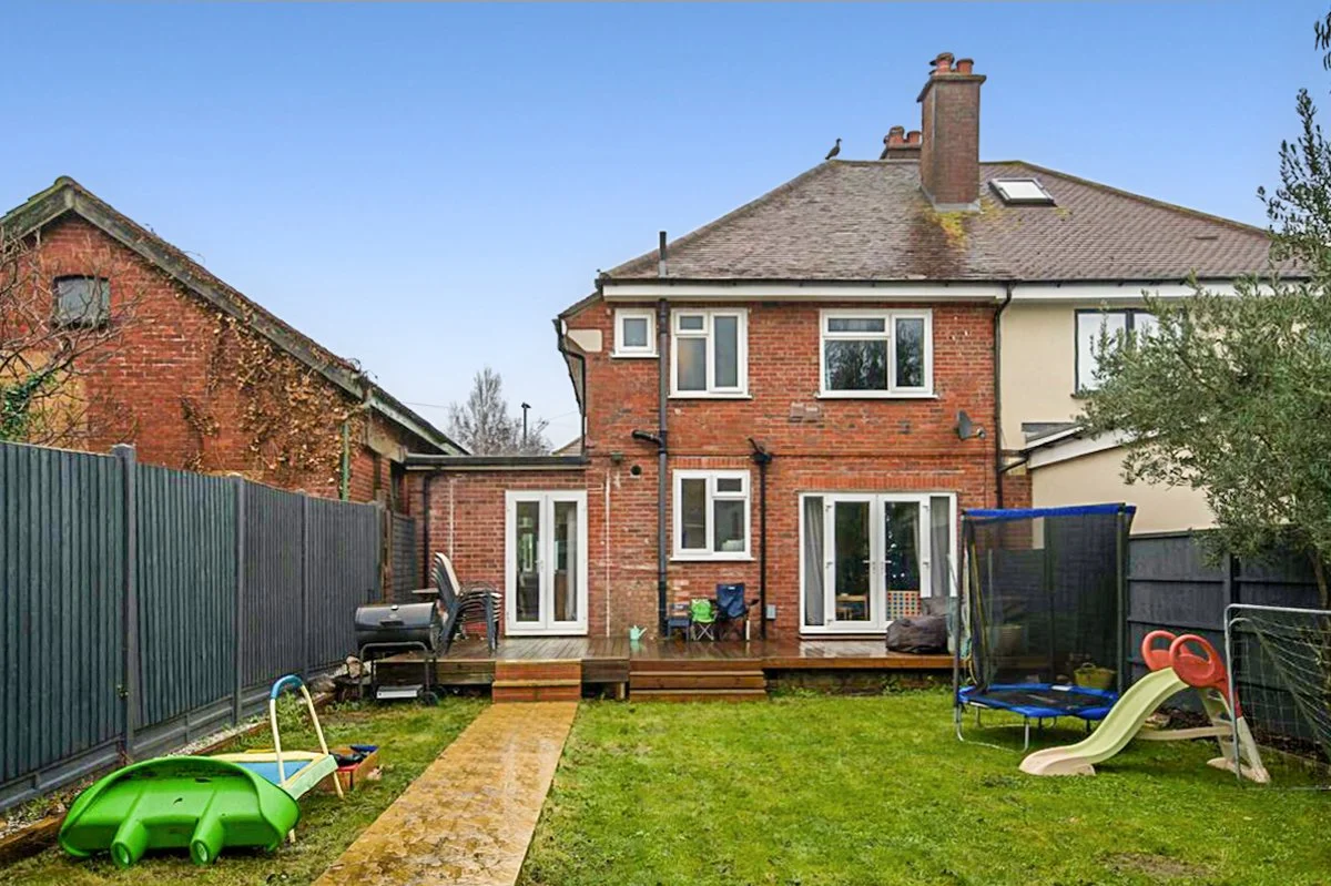 Rear view of a two-story brick house with a backyard, wooden deck, and children's play area including a slide and a trampoline, enclosed by a dark fence under a blue sky.
