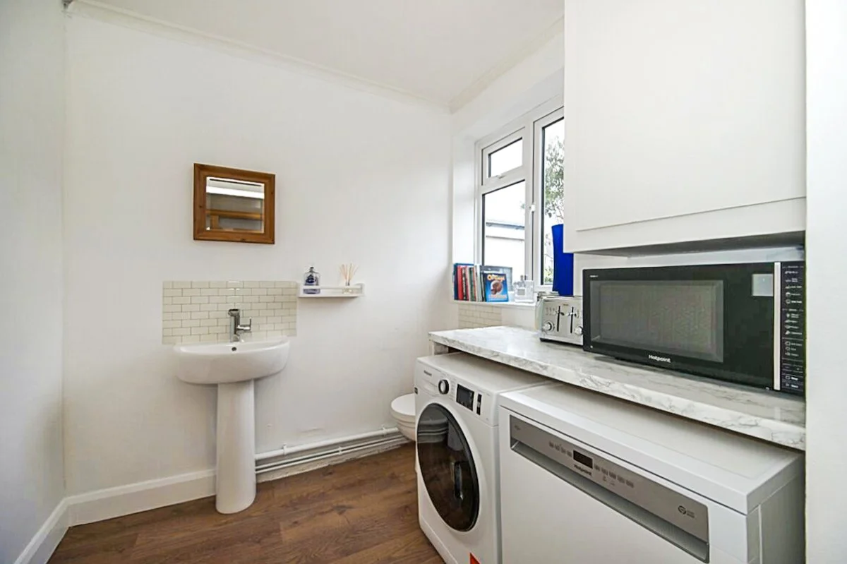 A small laundry room with a washing machine, microwave, and toaster on a marble countertop, a small sink on the left wall, and a window with outside view.