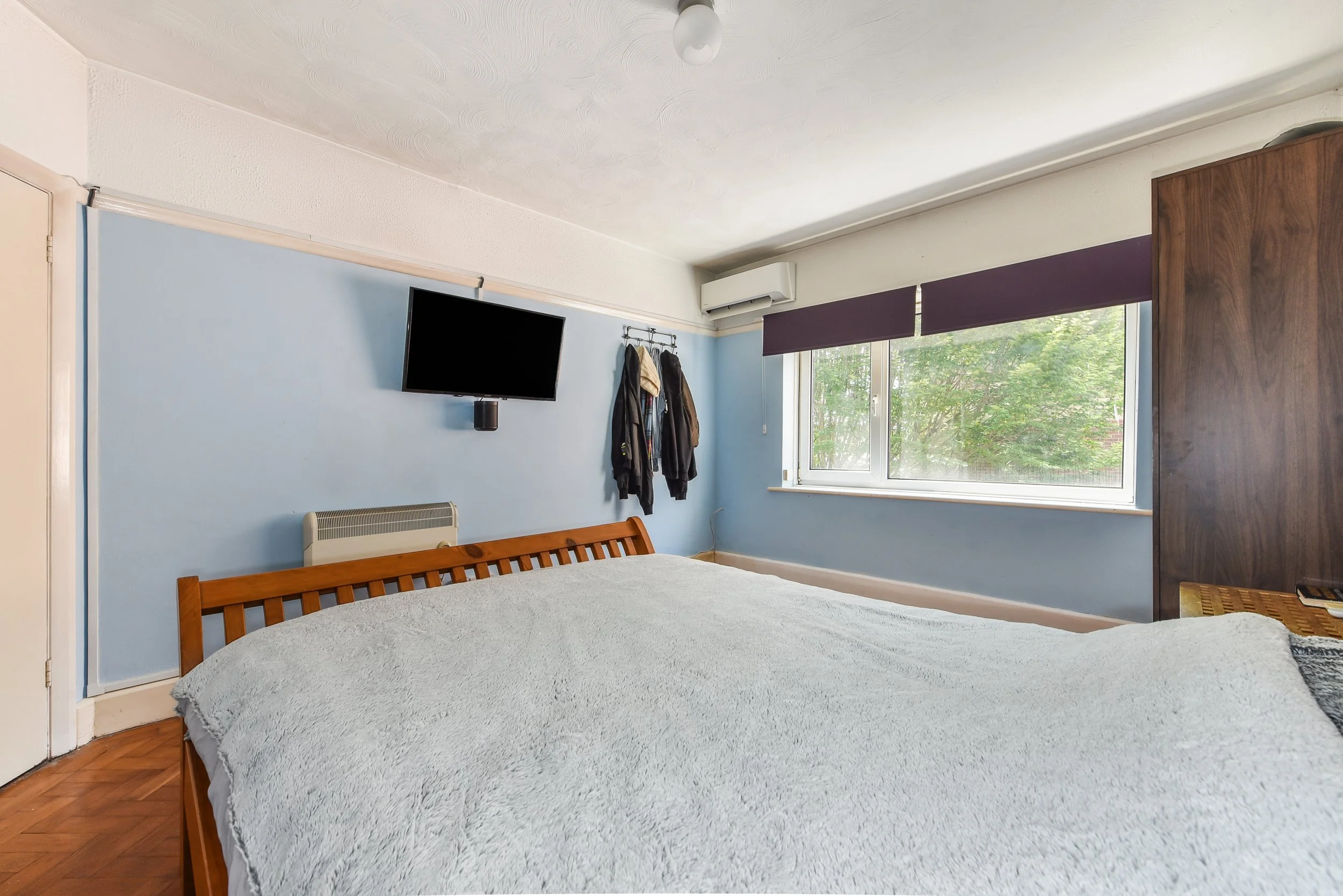 Bedroom with a wooden bed, a wall-mounted TV, large window with purple blinds, coatrack with clothes, and a dark wooden wardrobe.