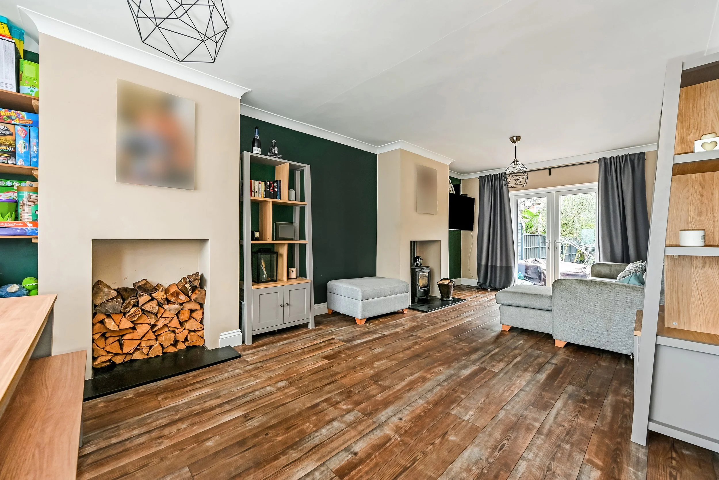 Living room with hardwood floors, beige and dark green walls, a fireplace with stacked wood, grey armchair and ottoman, bookshelf, flat-screen TV, large windows with grey curtains, and a sliding glass door leading outside.