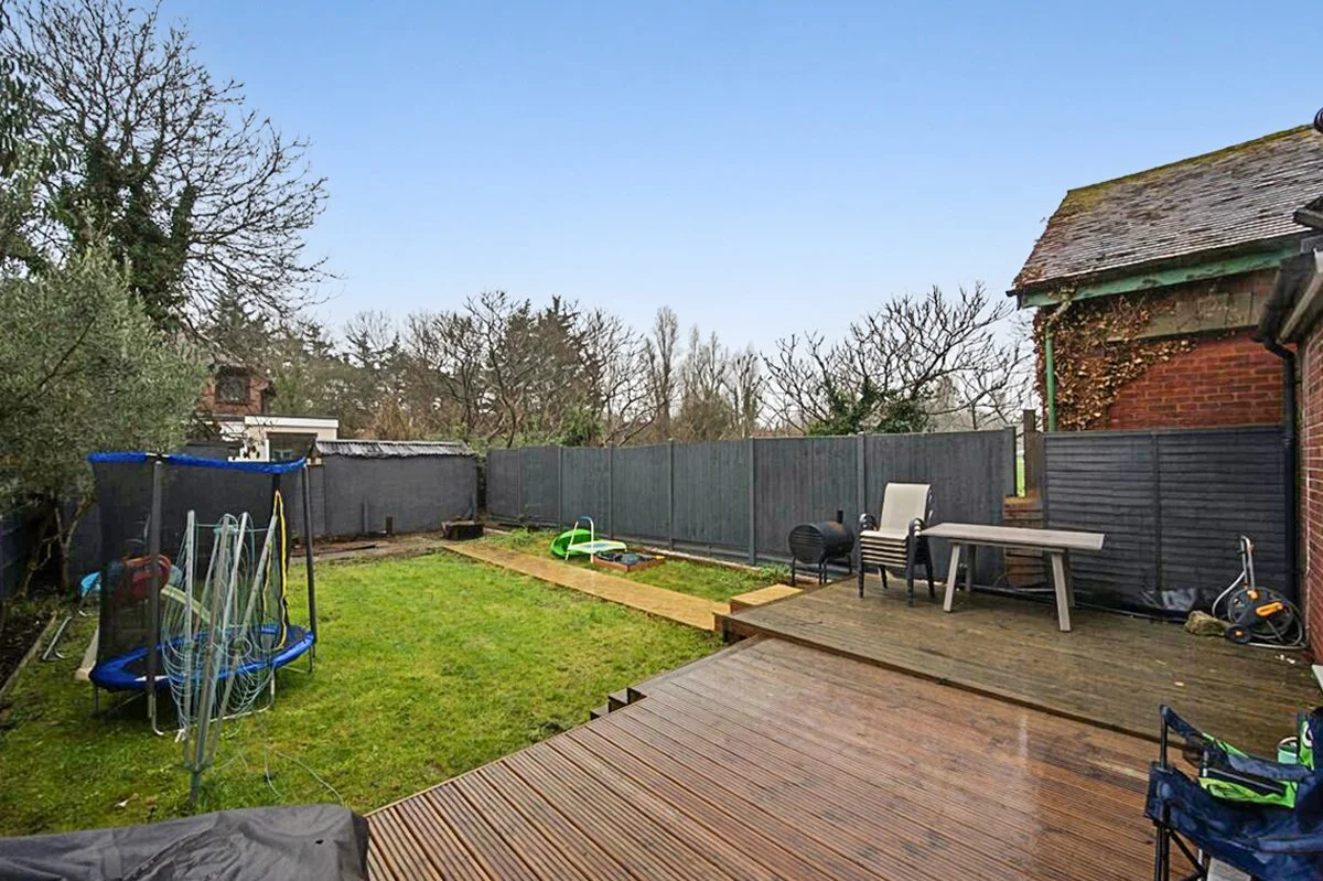 A backyard with a wooden deck, grass lawn, and a trampoline on the left. There are various garden beds and outdoor furniture, including chairs and a table, along the fence and on the deck. Trees and a blue sky are visible in the background.