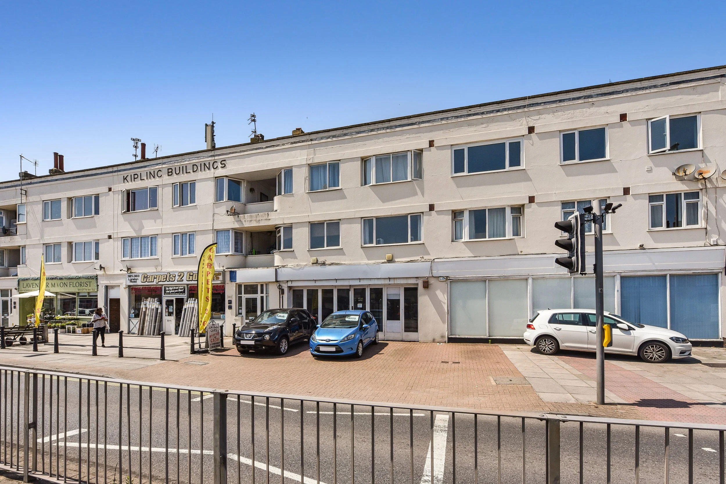 Street view of a white, three-story building with commercial shops on the ground floor, parking spaces in front, and a blue sky above.