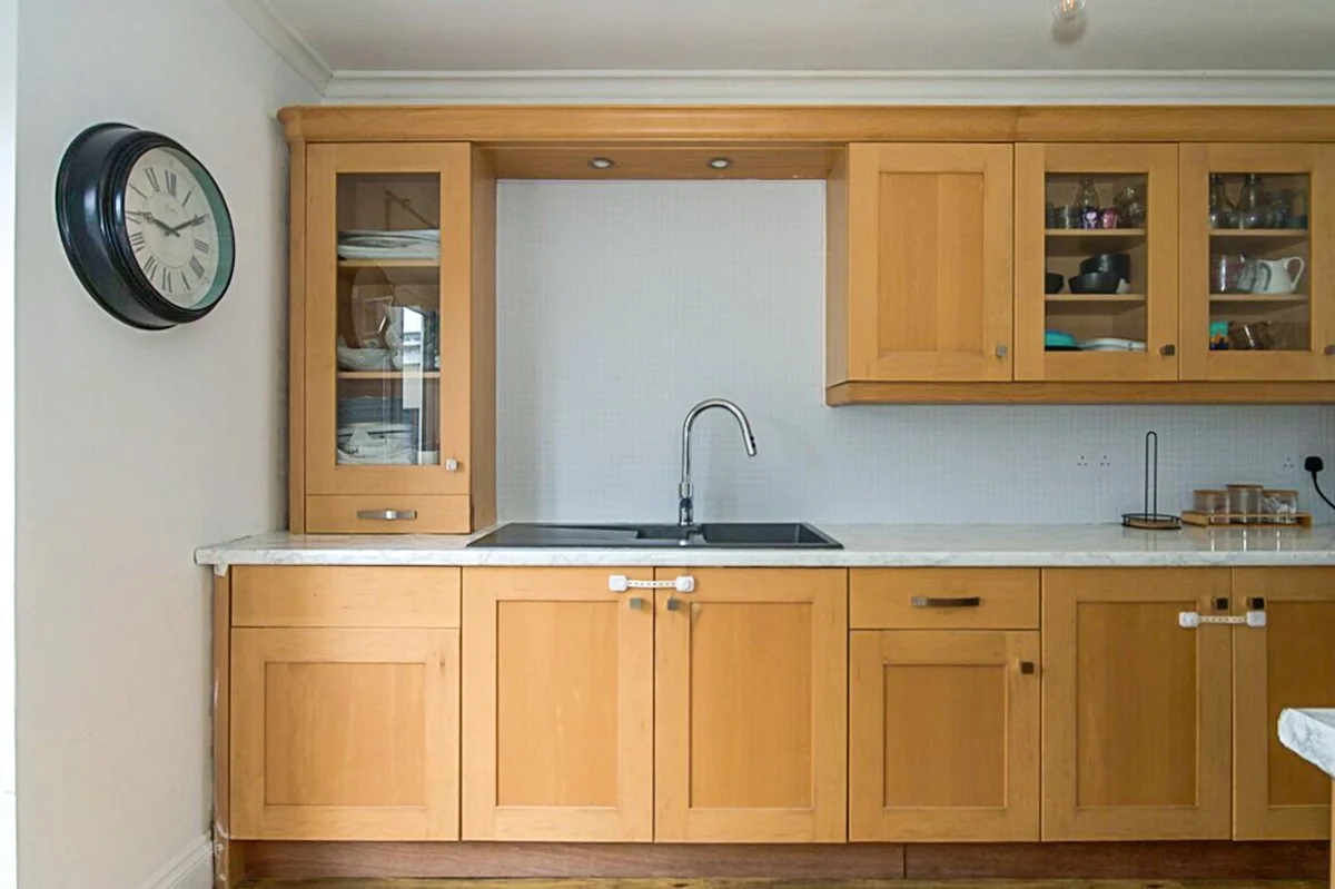Wooden kitchen cabinets with glass doors, a countertop with a sink, a wall clock, and some dishware inside the cabinets.