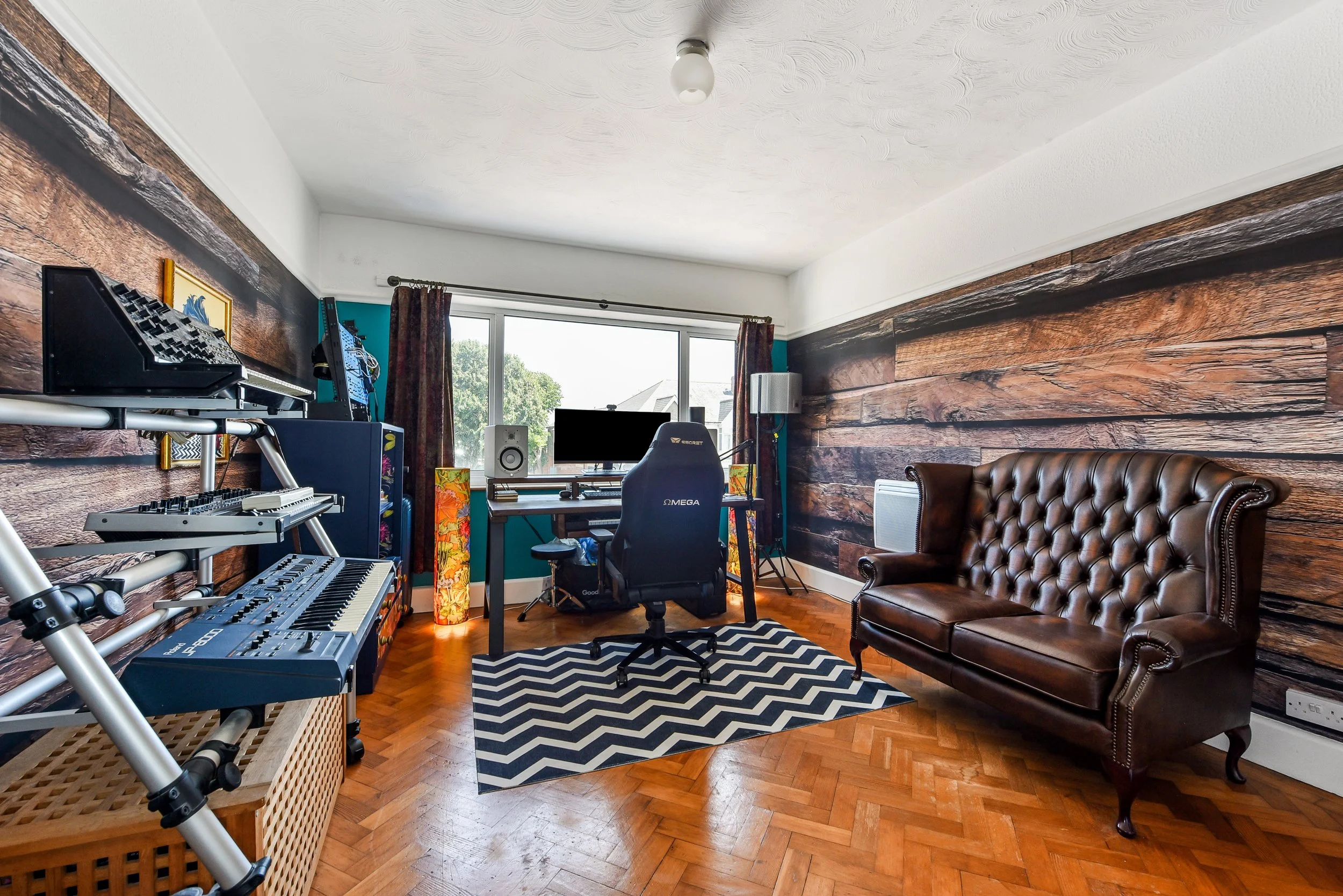 A music and recording studio room with wooden wall paneling, a desk with a computer and speakers, a black office chair, a brown leather sofa, and a large window with curtains.