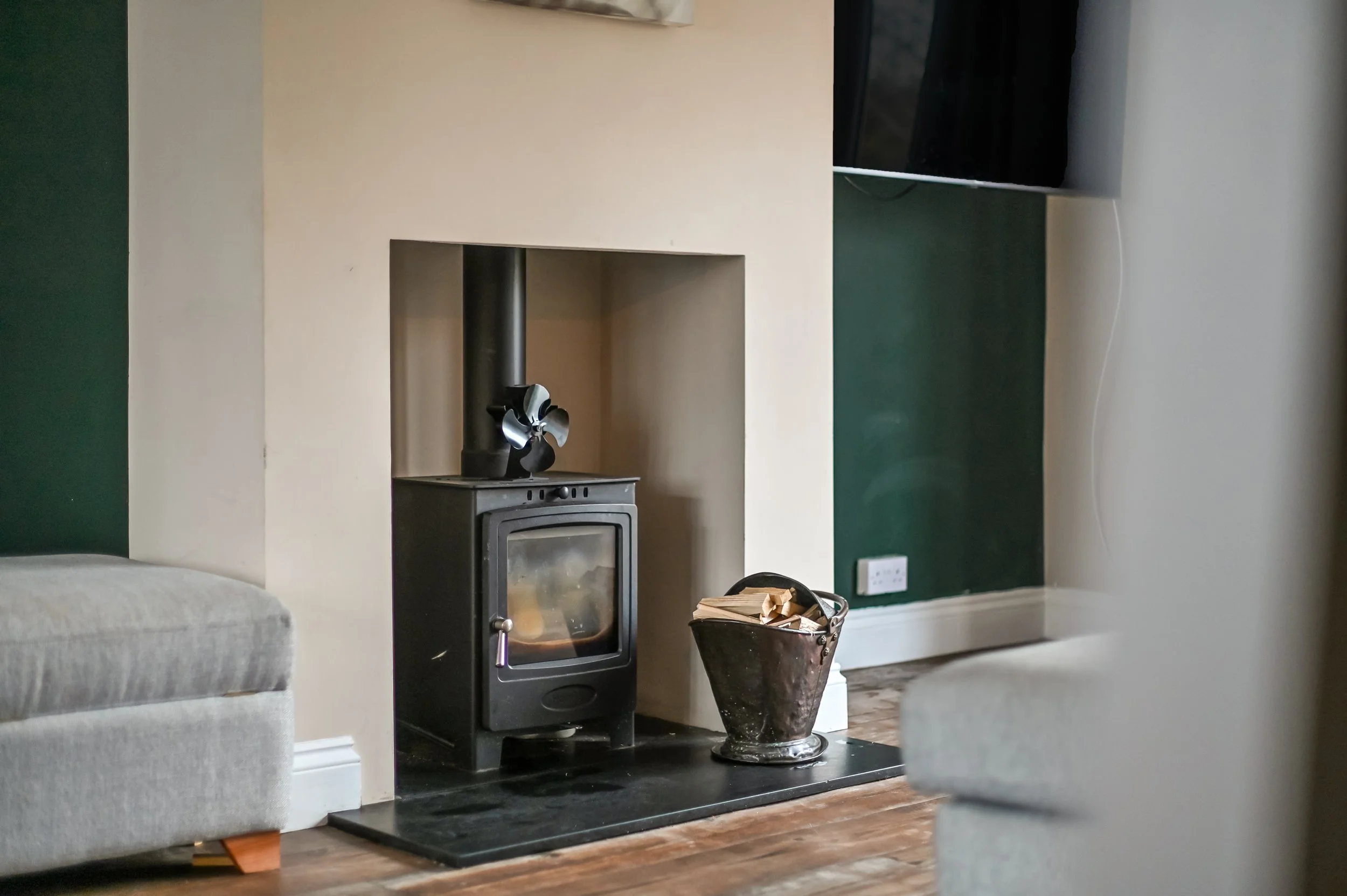 A cozy living room with a black wood stove on a black hearth, a bucket filled with firewood next to it, gray upholstered armchairs, and a flat-screen TV mounted on a dark green wall.