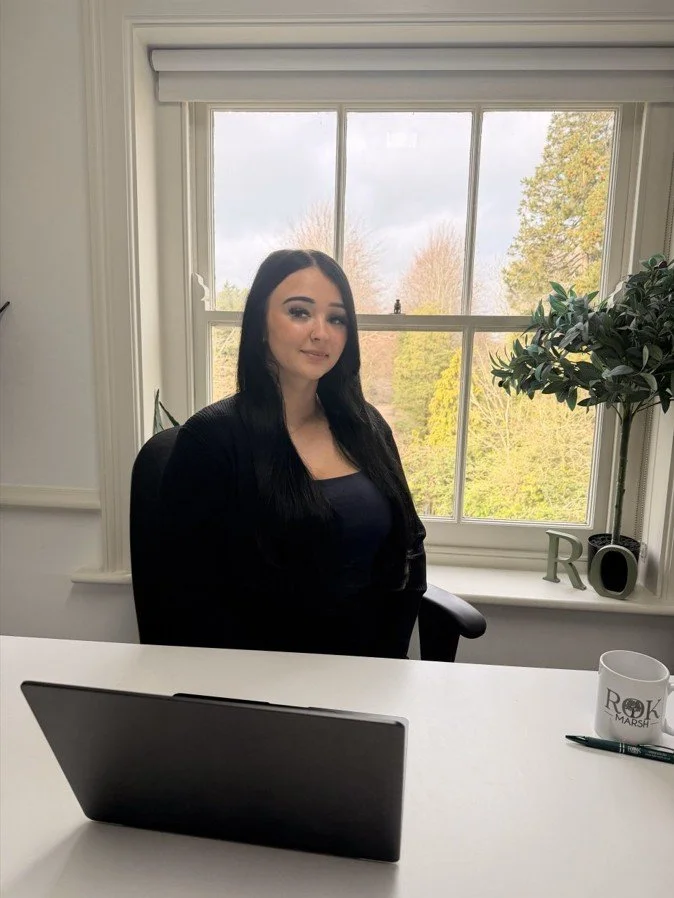 A woman with long dark hair sitting at a white desk in front of a laptop. There is a pen and a mug on the desk, and a potted plant on the windowsill behind her. The window shows a view of trees and a blue sky.