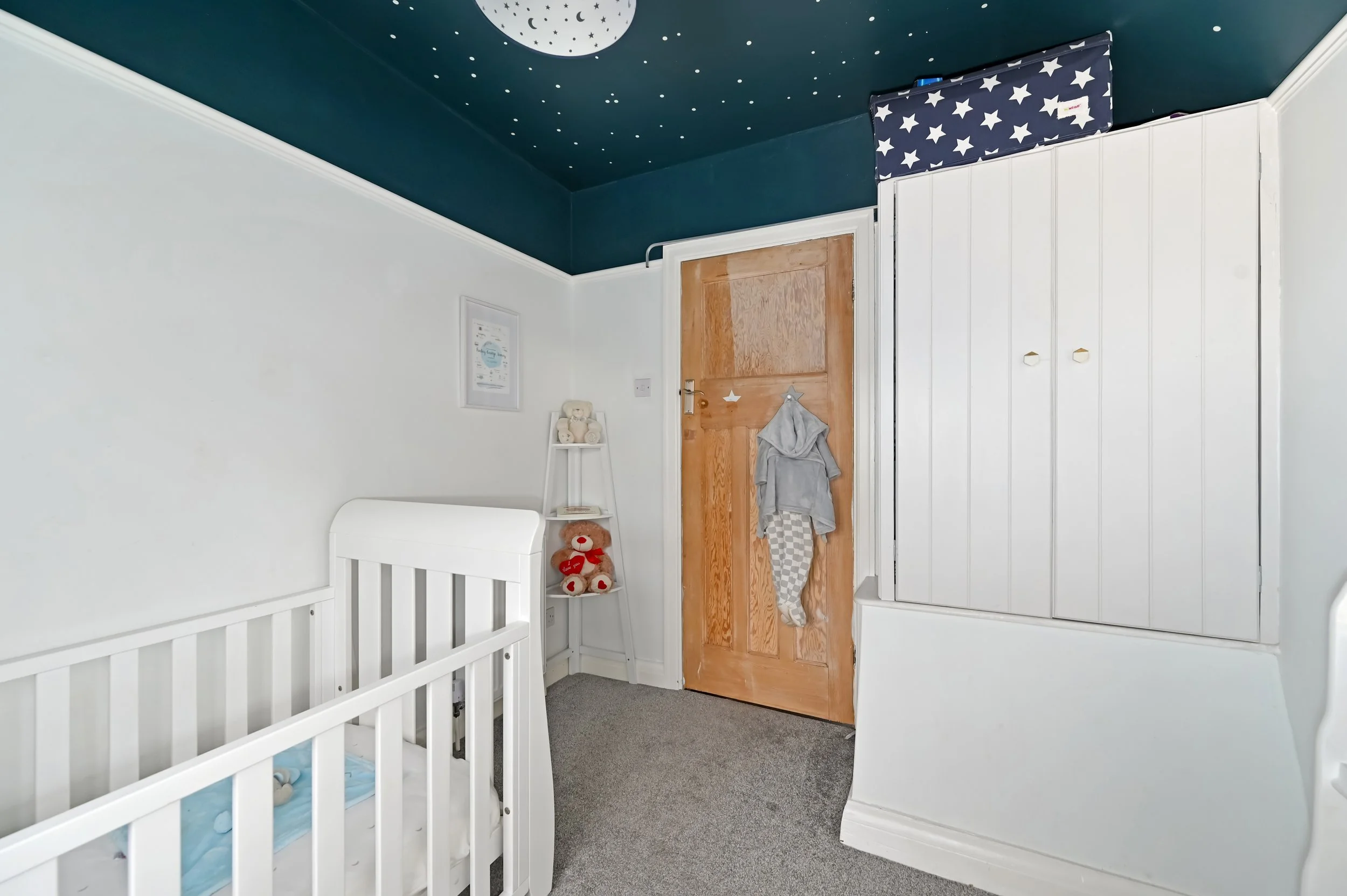 Baby crib, teddy bears on a small white shelf, and a closet in a child's nursery with a blue ceiling decorated with moon and stars.