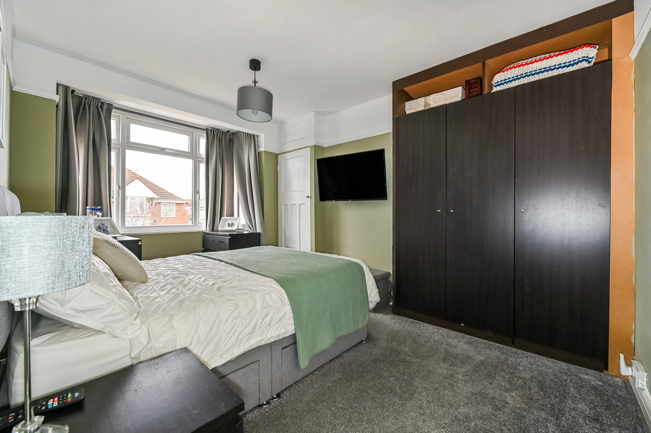 A bedroom with a large bed, gray and beige bedding, green throw blanket, nightstands with picture frames, a dresser with photo frames, a wall-mounted flat screen TV, a window with curtains, a ceiling light, and a large black wardrobe with stacked cus