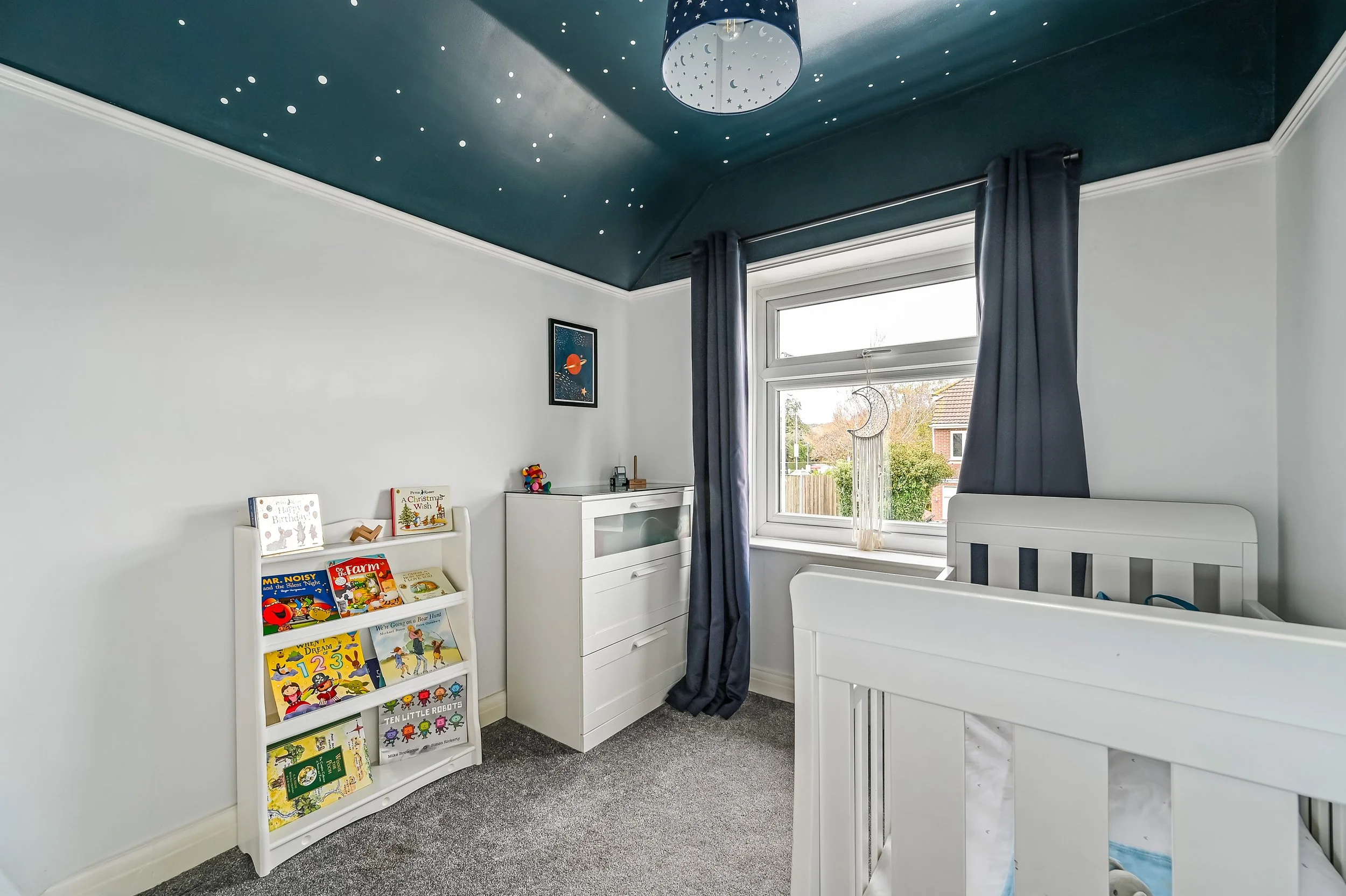 A child's bedroom with white walls and a dark blue ceiling decorated with star and moon patterns. The room has a window with dark curtains, a white crib, a white bookshelf filled with children's books, and a white dresser. There is a moon-shaped orna