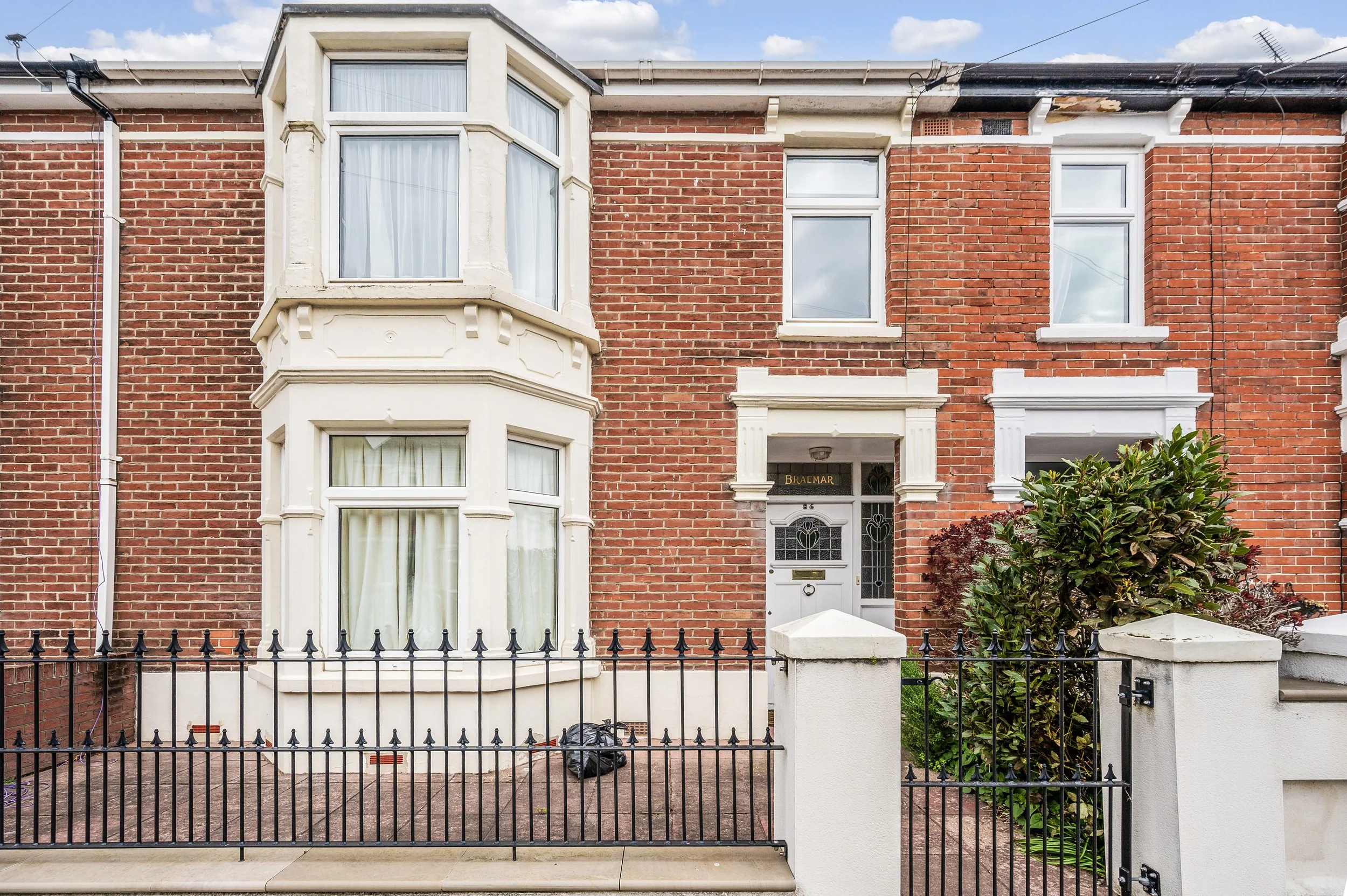 Red brick row house with bay windows, decorative white trim, and a black metal fence in front.