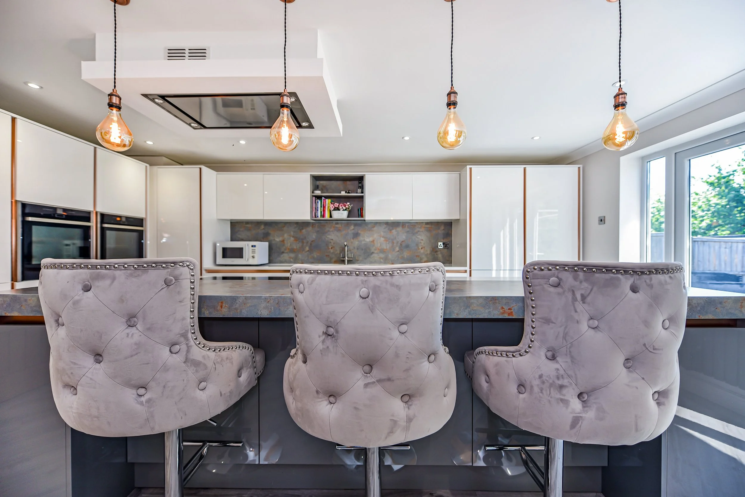 Modern kitchen with a large island, three plush beige bar stools, white cabinetry, and exposed light bulbs hanging from the ceiling.