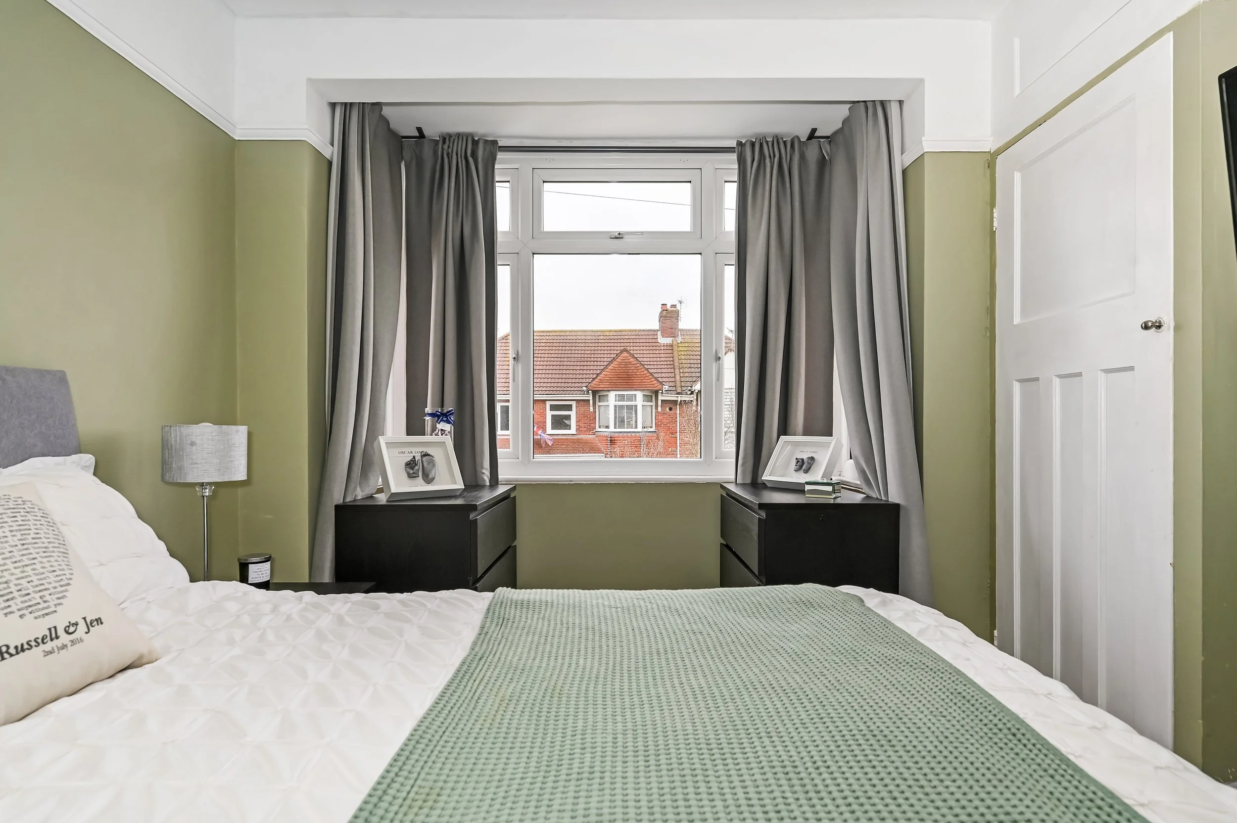 Interior of a bedroom with a bed, two black nightstands, a large window with curtains, and decorative items on the nightstands.