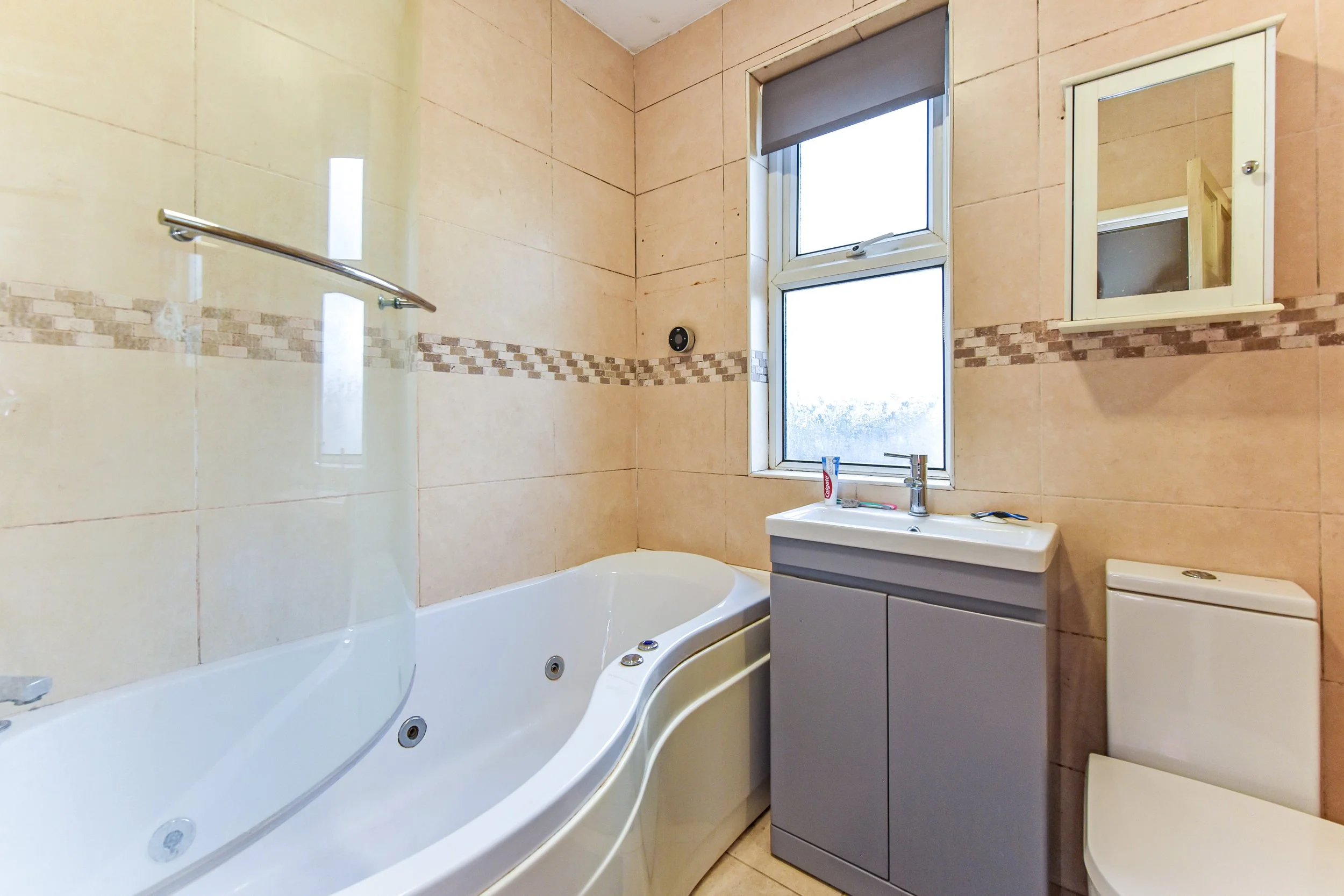 A bathroom with beige tiled walls, a white bathtub, a gray vanity with sink, a mirror cabinet, a toilet, and a window with a roller blind.