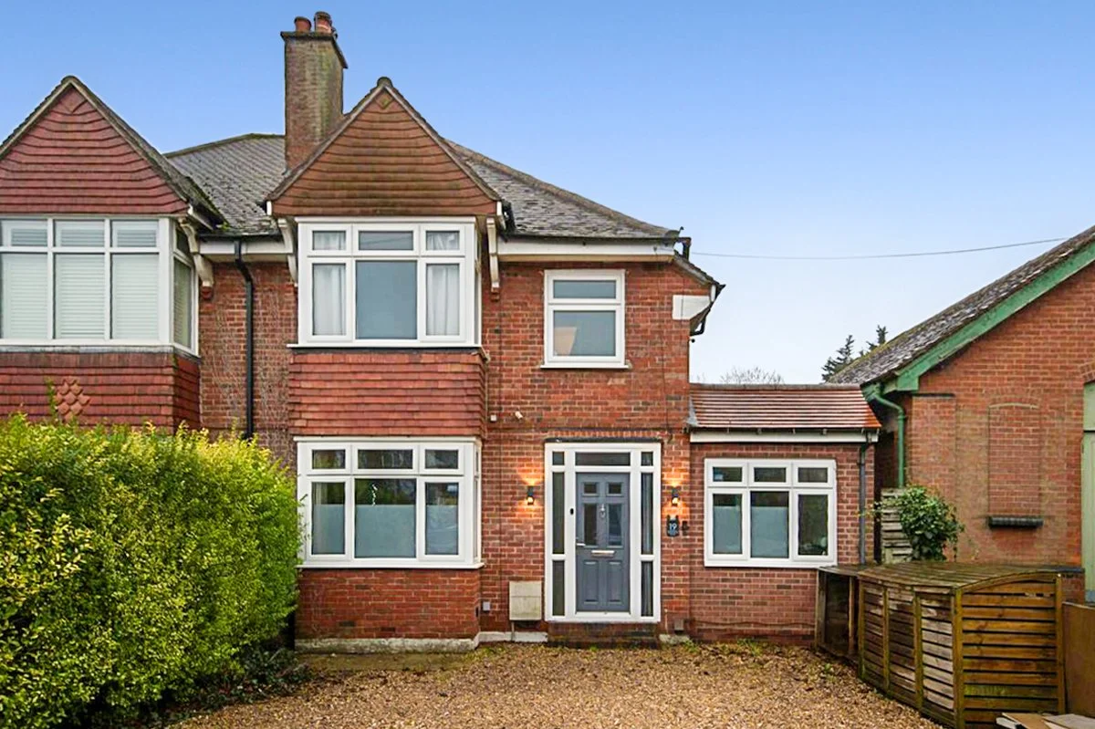 A two-story brick house with a gray front door, surrounded by multiple windows, a shrubbery on the left, and a small gravel driveway in front.