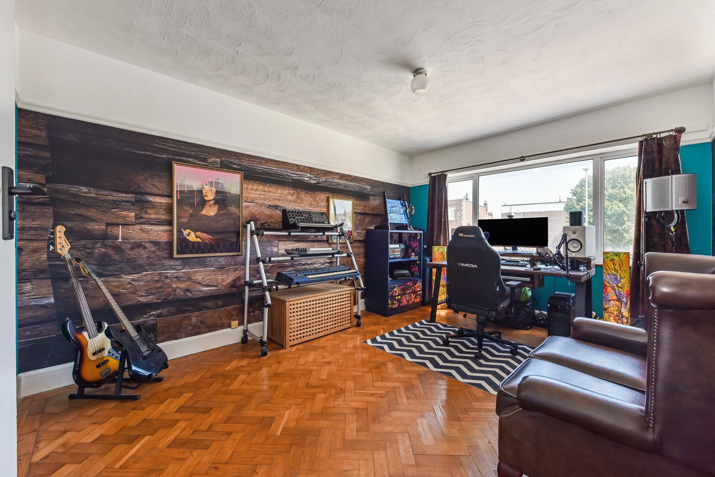A music production and gaming room with guitars, a computer, speakers, and musical equipment, featuring a wooden accent wall and a large window.