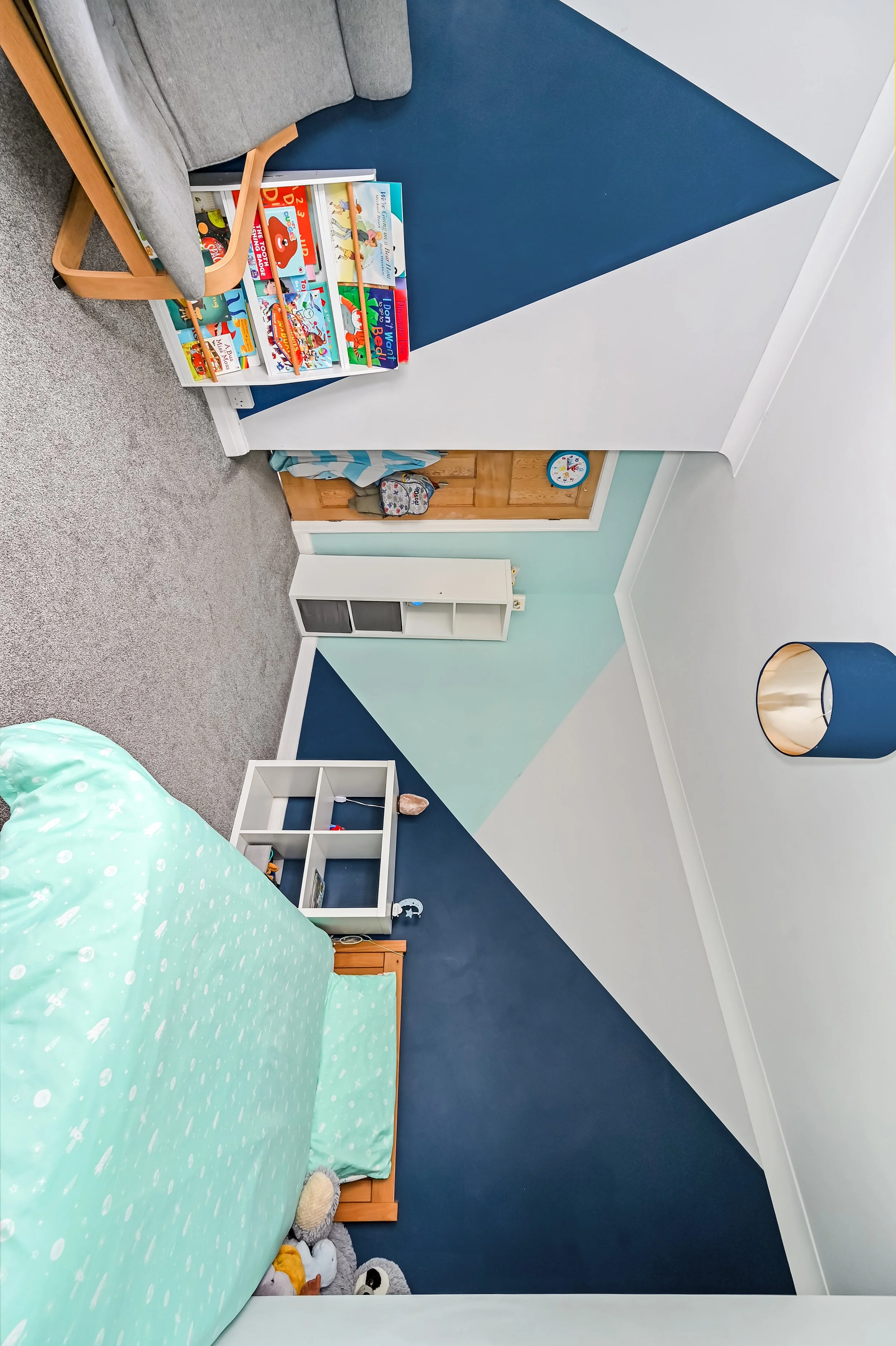 Child's bedroom with a bed covered in a light blue duvet, a white bookshelf, a small white cabinet, and colorful geometric wall paint in shades of blue, white, and green.