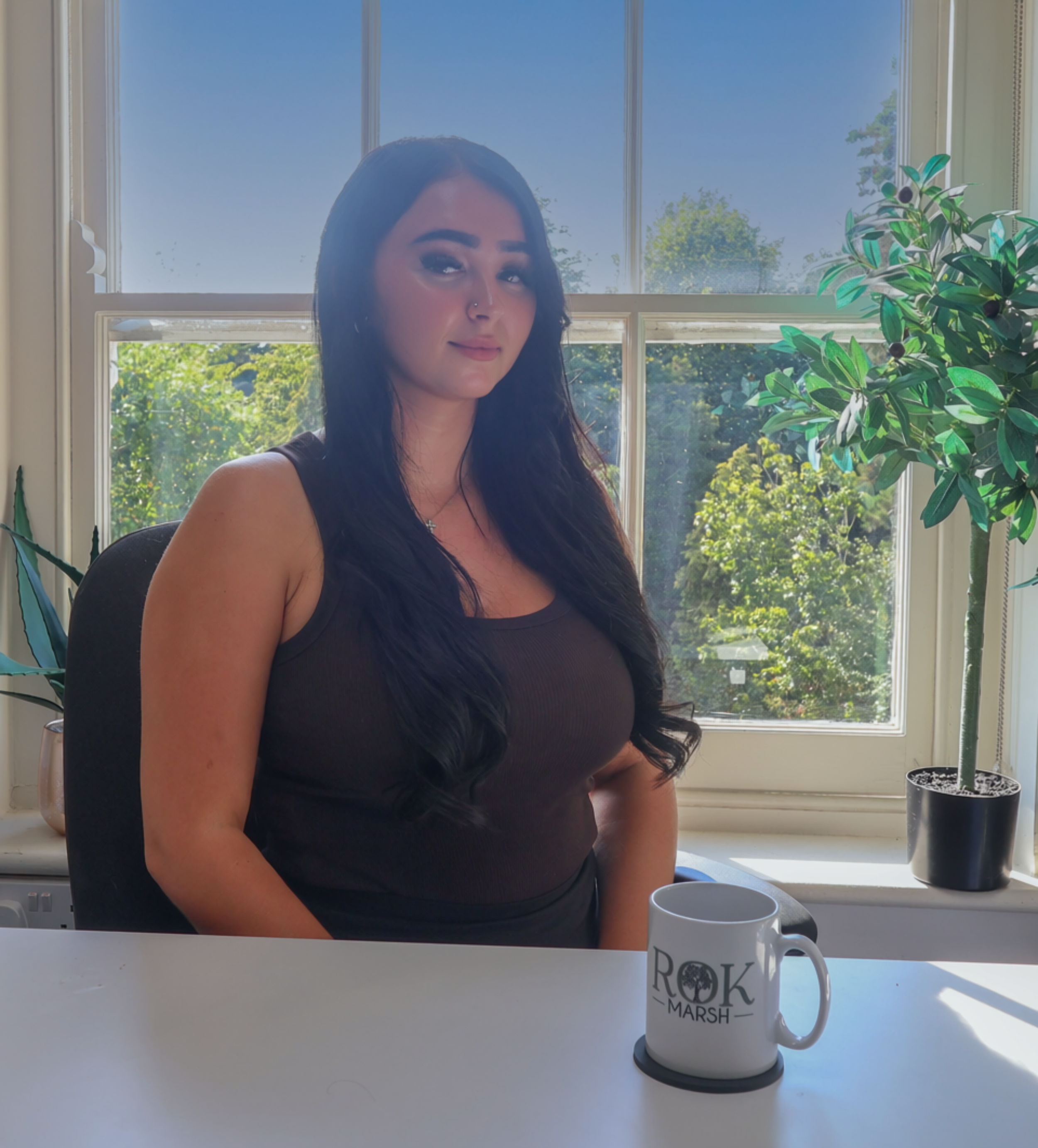 A woman with long dark hair sitting at a white desk in front of a laptop. There is a pen and a mug on the desk, and a potted plant on the windowsill behind her. The window shows a view of trees and a blue sky.