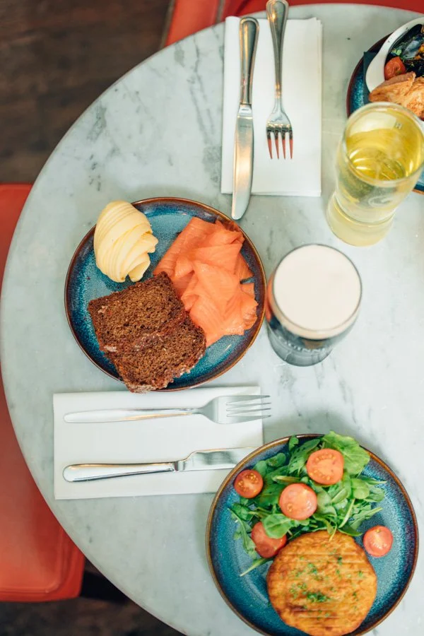 A breakfast table with two plates of food, two glasses of drinks, and cutlery. One plate has slices of smoked salmon and a slice of dark bread, the other has a green salad with cherry tomatoes and a savory pancake.