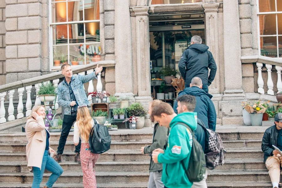 Group of people standing and sitting on the steps of a building with potted plants for sale outside.