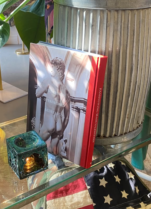 A book titled 'Undressed Overdressed Redressed' leaning against a large metallic planter with vertical ridges on a glass table. Next to the book is a decorative teal-colored candle holder with a lit candle inside. The background includes a large green plant with broad leaves.