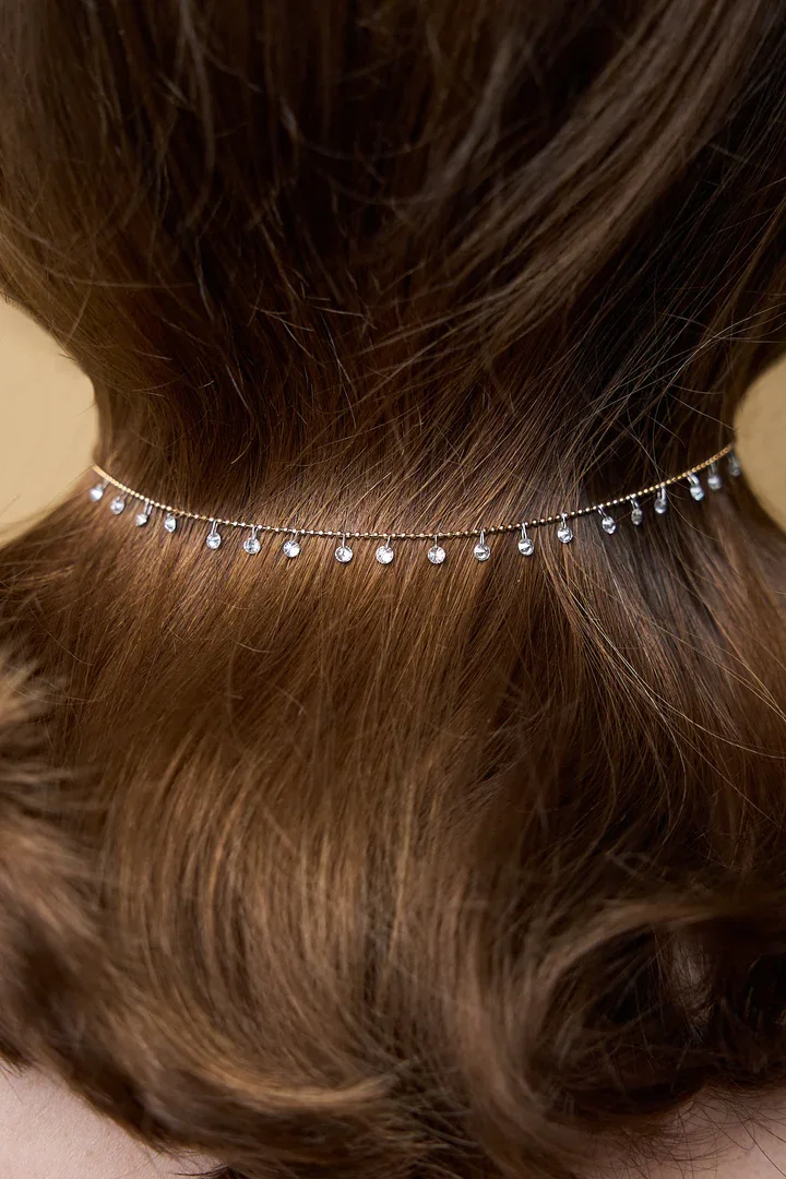 Close-up of a woman's neck wearing a delicate jewelry piece with small dangling pearls or beads, with her wavy brown hair partially covering the jewelry.