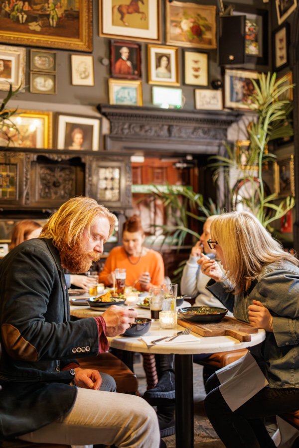 A group of people dining in a cozy restaurant with vintage decor, including framed artwork on the wall and large indoor plants.