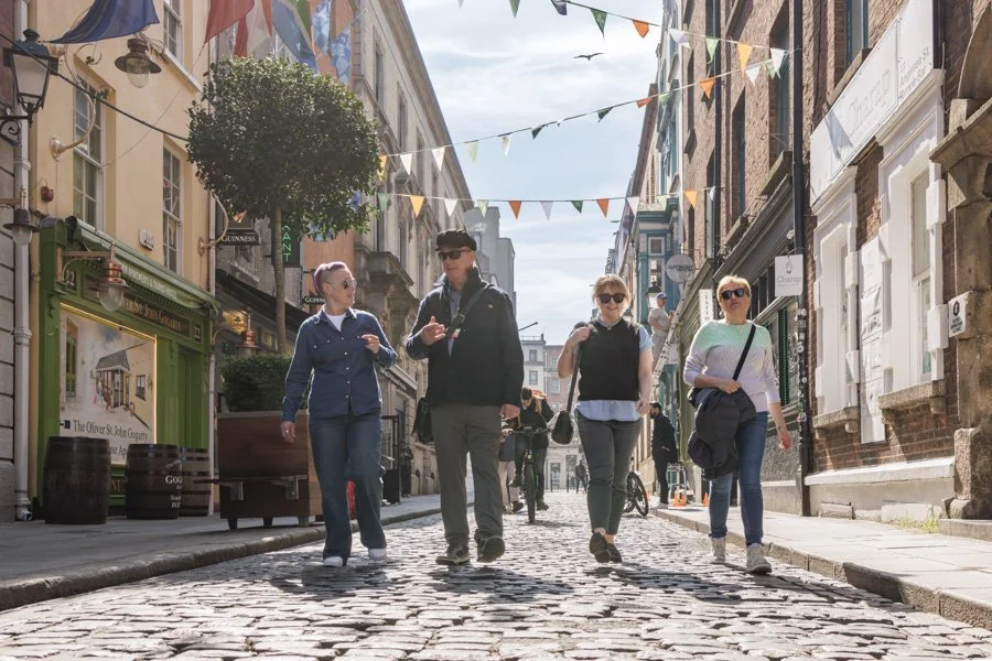 Four people walking on a cobblestone street in a city with colorful bunting flags overhead, shops and buildings lining the street, and a clear sky.