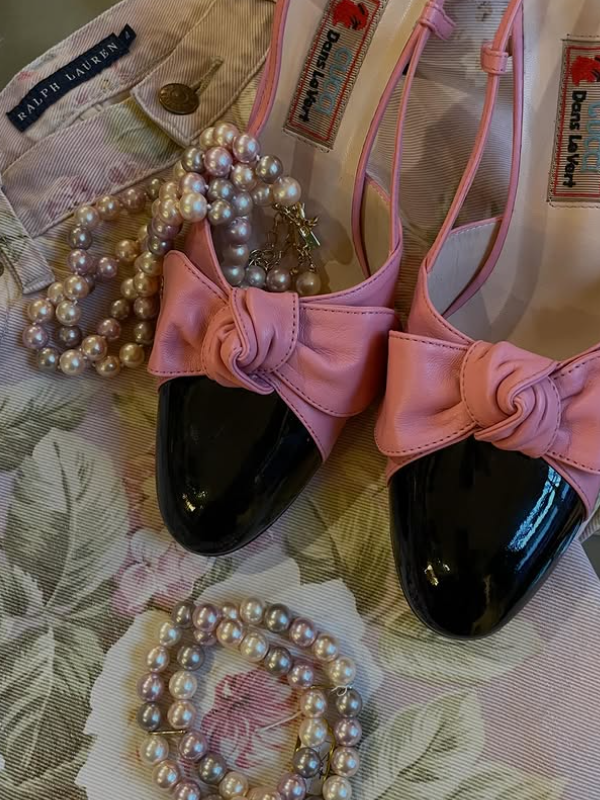 Pink and black shoes with bows, pearl necklaces, and a Ralph Lauren bag on a floral tablecloth.