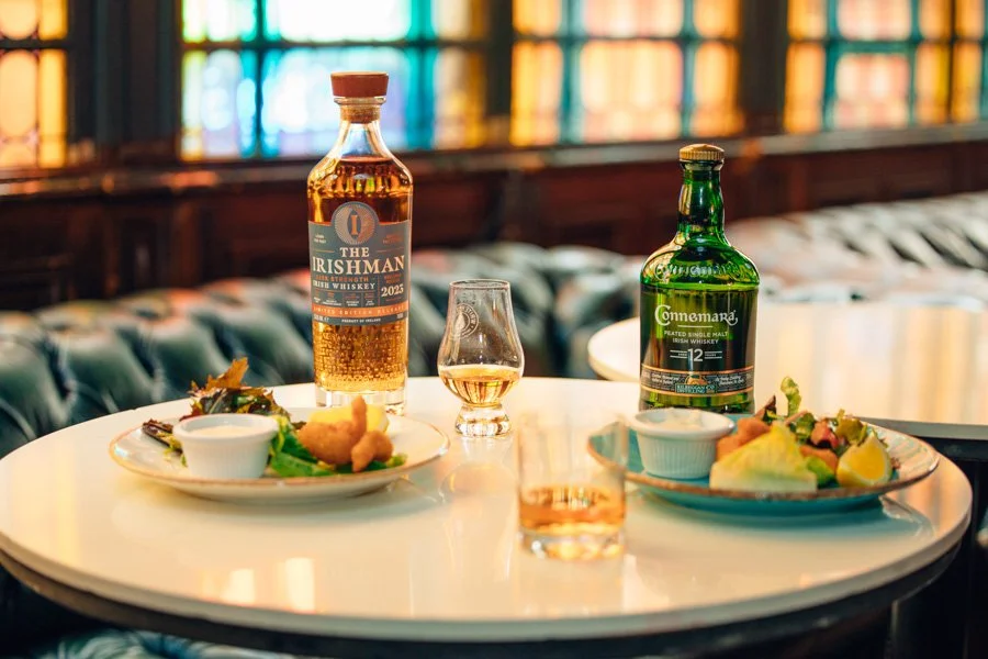 A round white table set with two plates of food, two bottles of whiskey, two glasses of whiskey, and a small glass of water. The background features colorful stained glass windows.