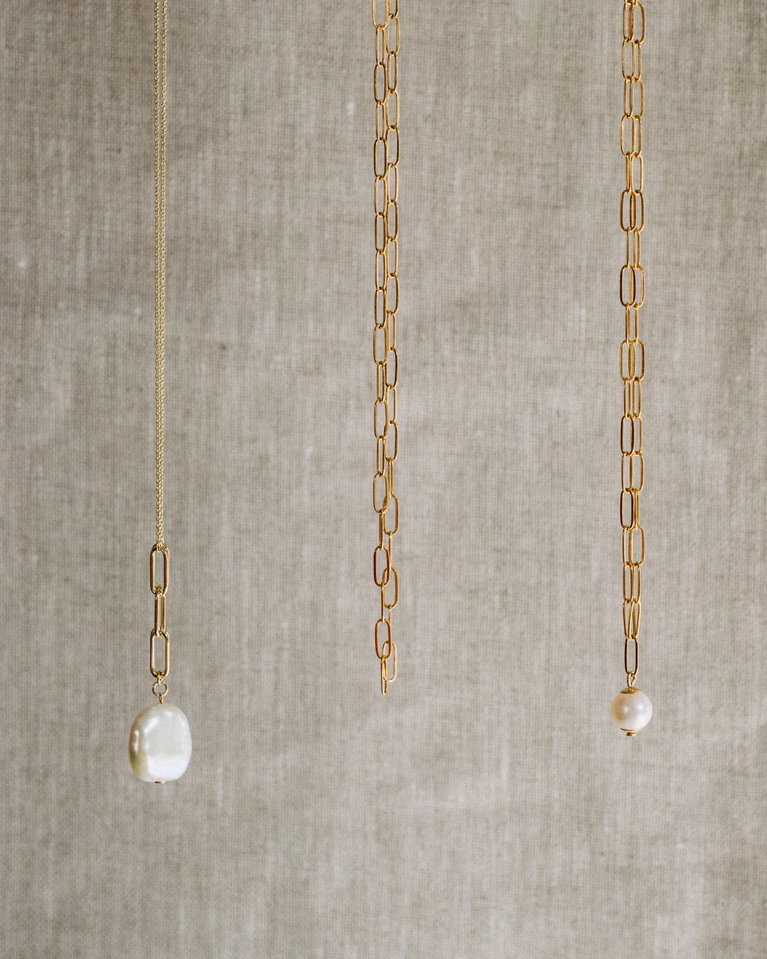 Three gold necklaces with pearl pendants hanging against a beige background.
