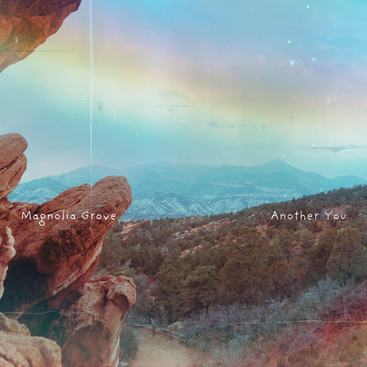 A landscape photo showing rocky formations in the foreground, trees and rolling hills in the midground, and mountain ranges in the background under a sky with a rainbow and clouds. Text labels indicate areas named 'Magnolia Grove' and 'Another You'.