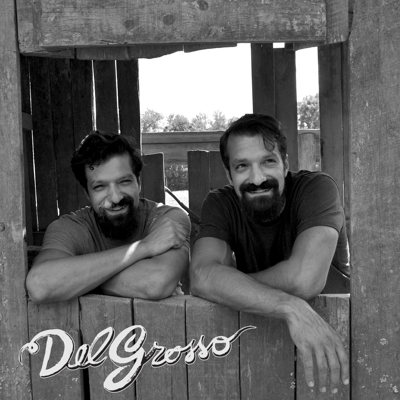A black-and-white photo of two men with beards and dark hair, leaning on a wooden fence, smiling. The photo appears to be taken outdoors.