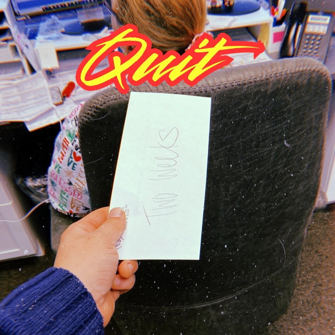 A person holds a piece of paper with the text 'I Love You' written on it in front of an office chair. The chair has a label or sticker with colorful text and icons, and the background shows a cluttered desk with office supplies. A large red and yello