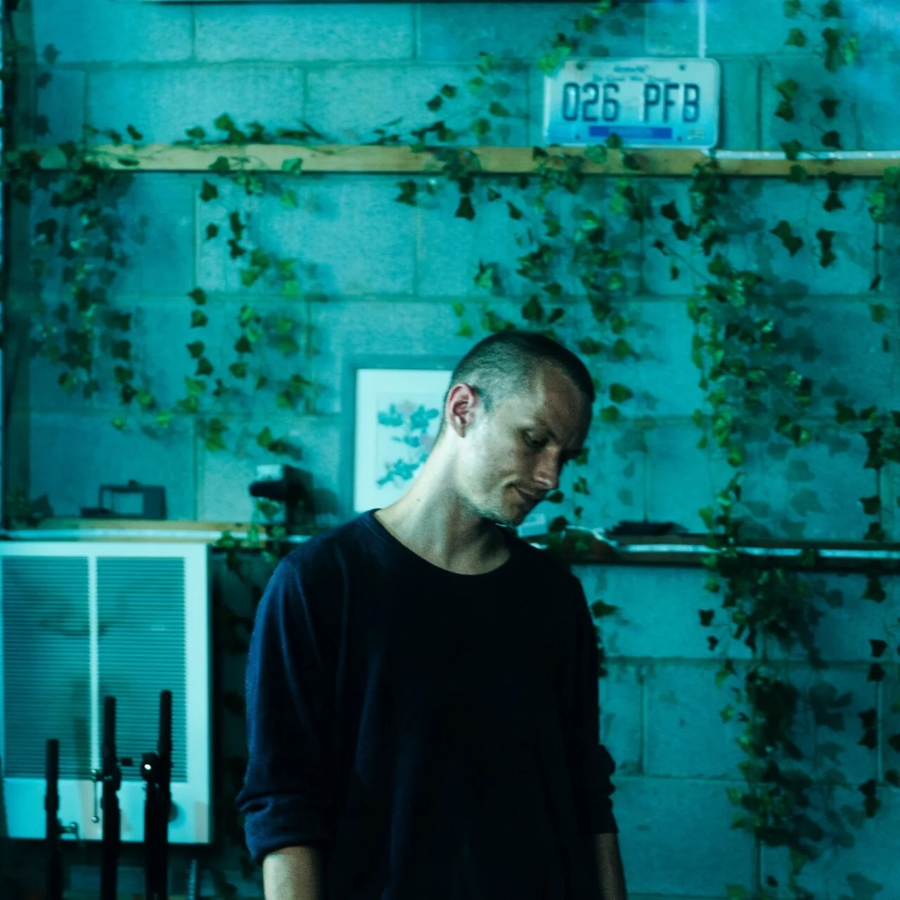 A young man with a shaved head wearing a dark long-sleeve shirt stands in front of a gray brick wall decorated with ivy. There is a framed picture and a license plate on the wall, with some electronic equipment and wires below.