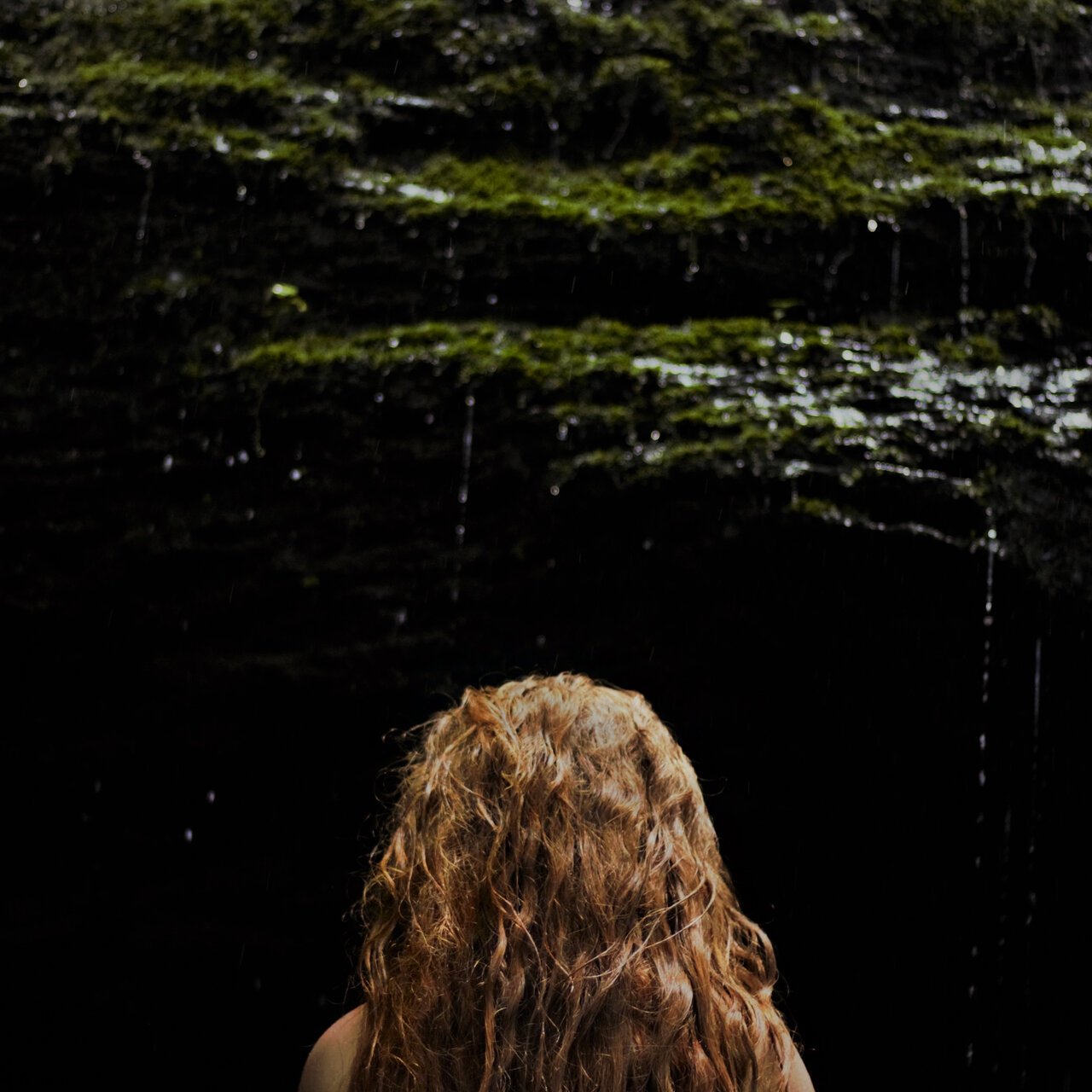 Back view of a person with curly red hair looking up at a dark, moss-covered cave ceiling with water dripping from above.