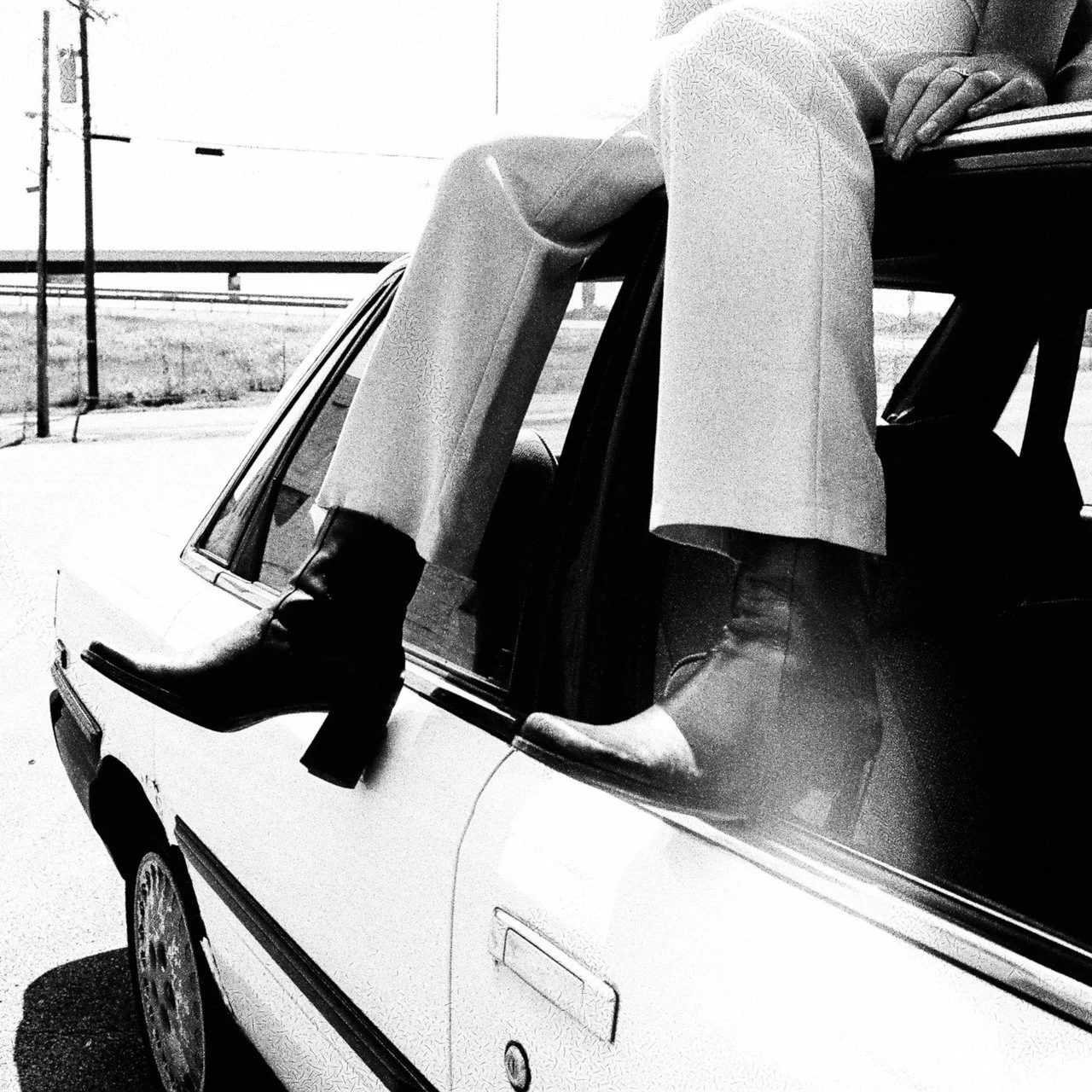 A person sitting on the roof of a vintage car with one foot hanging out of the window, wearing heeled boots and light-colored clothing, in a black-and-white photo.