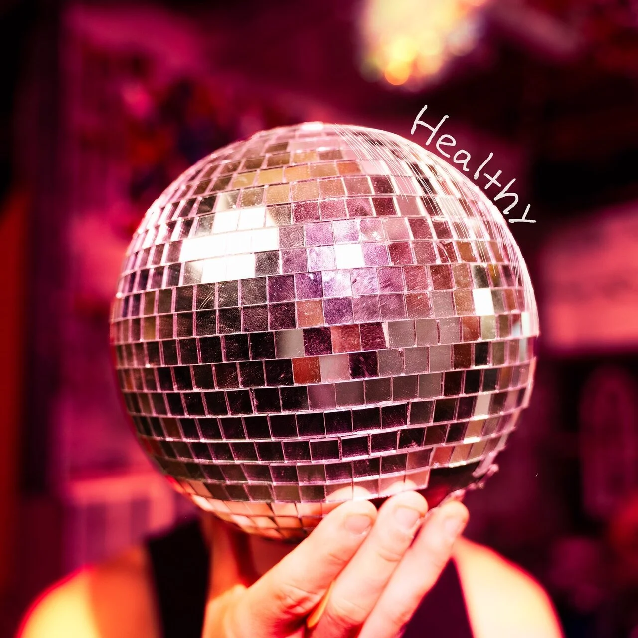 Person holding a disco ball mask with the word 'Healthy' written on it, with a pink and purple background.